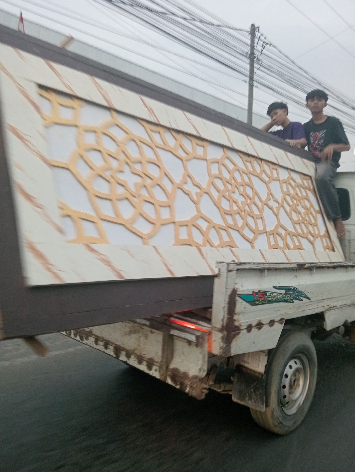 The image depicts a light-duty truck carrying a large, decorative panel with an intricate golden geometric pattern on a light-colored background, overlaid with reddish-brown streaks. Two young males, likely teenagers, are positioned on top of the panel. One is wearing a dark t-shirt with a graphic design and jeans, while the other wears a purple t-shirt. The truck is in motion on a paved road, likely in Pringsewu, Indonesia, indicated by the presence of overhead electrical wires and a utility pole typical of urban or semi-urban environments in the region. The truck has a "SUPER CARGO" logo on its side. The overall scene is slightly blurred, suggesting movement.