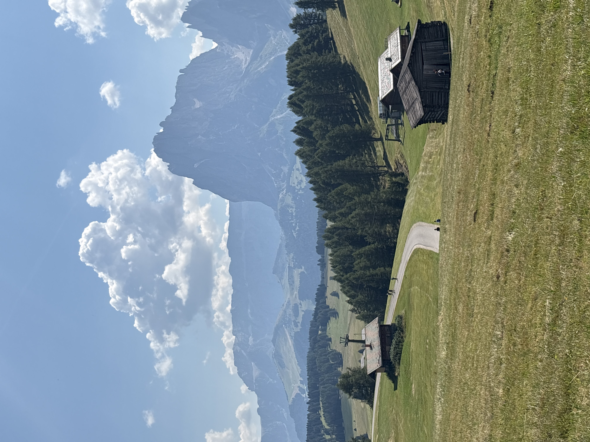 The image depicts a scenic mountain landscape in Kastelruth - Castelrotto, Italy. In the foreground, a grassy meadow slopes gently upwards. A winding paved road traverses the middle ground, leading past a small, rustic wooden cabin with a red-tiled roof. A ski lift structure is visible next to the cabin. Further along the road, several figures, appearing to be people walking or cycling, are present. To the right of the road, another, older, dark wooden shed stands. The background is dominated by a dense forest of evergreen trees, beyond which are layered mountain ranges that recede into a hazy blue distance. The sky above is a clear blue with scattered, puffy white clouds, and a prominent, jagged mountain peak is silhouetted against the sky in the far background. The lighting suggests a bright, sunny day.