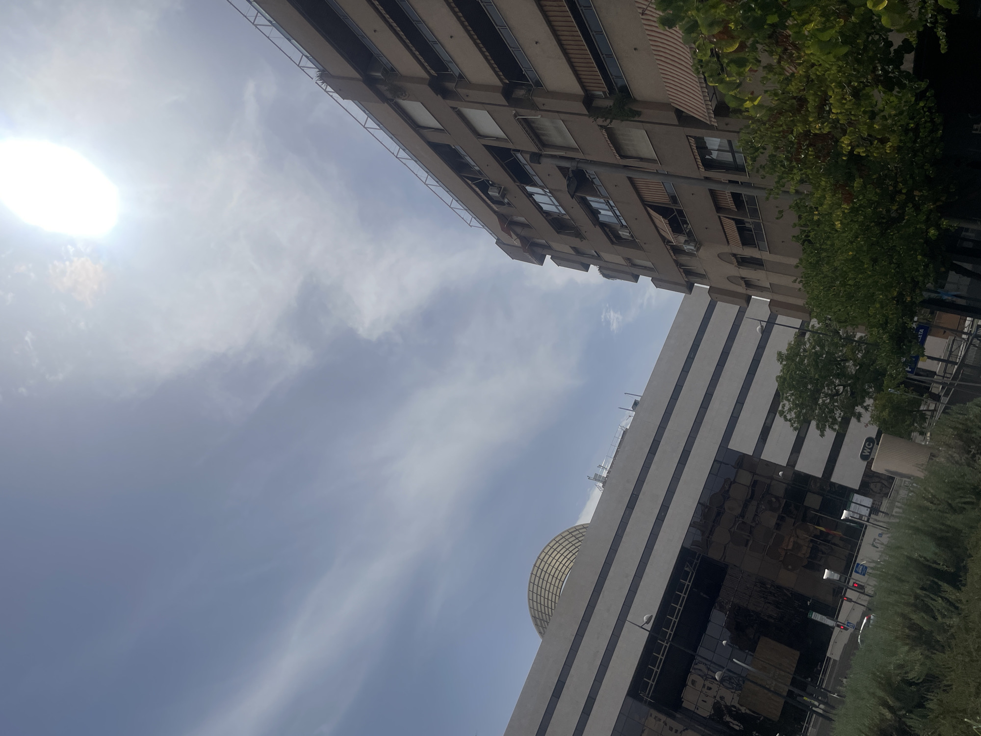 The image shows a bright, sunny day in Granada, Spain, with a clear blue sky and some wispy clouds. The sun is prominently visible in the upper right corner, creating a lens flare effect. In the foreground, to the left, is a modern building with a large glass facade, reflecting the sky and surrounding structures. On the right, an older, multi-story apartment building with a brown facade is visible, featuring balconies with awnings and air conditioning units. A rounded, metallic dome-like structure can be seen atop the modern building. In the lower part of the image, there is dense green foliage, suggesting a park or landscaped area. The scene appears to be an urban environment, likely a commercial or residential area.