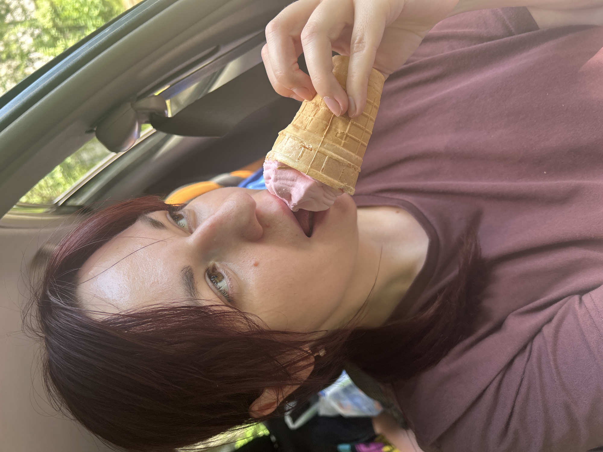 A woman is eating an ice cream cone inside a car. The woman has dark red hair and is wearing a maroon t-shirt. The ice cream is pink and appears to be strawberry flavored. The car interior is visible, including the ceiling and a portion of the passenger seat. The setting appears to be outdoors, with trees and greenery visible through the car window. The sunlight suggests it is daytime, and the weather appears to be warm and pleasant. The woman's expression is relaxed and she seems to be enjoying her ice cream. There is no visible text in the image.