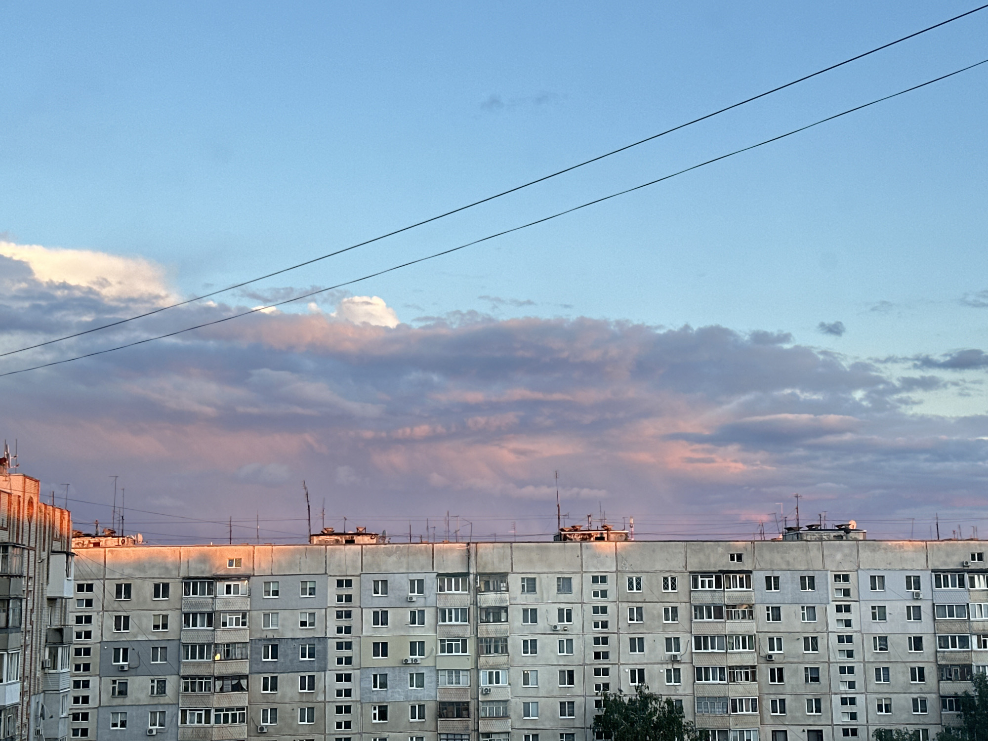 The image shows a multi-story residential building in an urban setting. The building is characterized by a repetitive pattern of windows and balconies. Electrical wires run across the top portion of the frame, juxtaposed against a sky filled with clouds tinged with pink hues, suggesting either sunrise or sunset. The building's facade appears to be constructed from concrete or a similar gray material. The overall scene captures an ordinary urban landscape under a colorful sky.