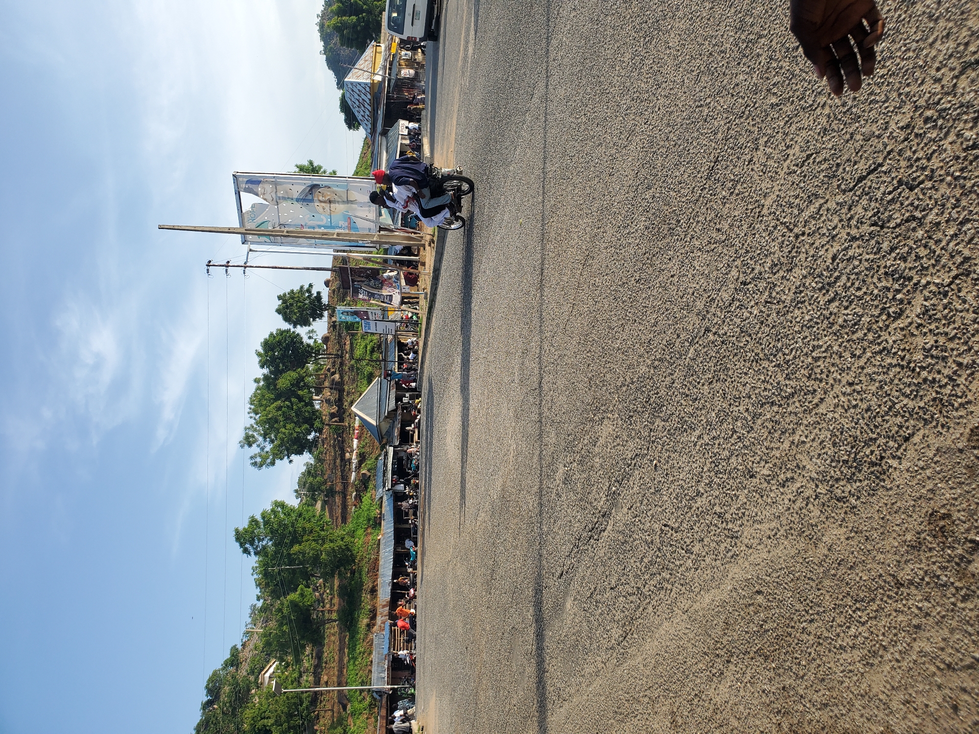 The image depicts a street scene in the city of Ampan, Nigeria. In the foreground, a paved road surface is visible. In the middle ground, a group of market stalls with corrugated metal roofs are lined up, with various people walking and standing around them. A few motorcycles and a white van are parked in the vicinity. A prominent banner, likely a political advertisement, stands on a pole to the right of the market stalls. In the background, a hilly terrain with lush green vegetation and scattered trees can be seen, with a few houses or structures nestled on the slopes. The sky is clear and blue with some scattered clouds. Two individuals are riding a motorcycle in the mid-ground, moving towards the right of the frame. A hand is visible in the bottom right corner of the image.