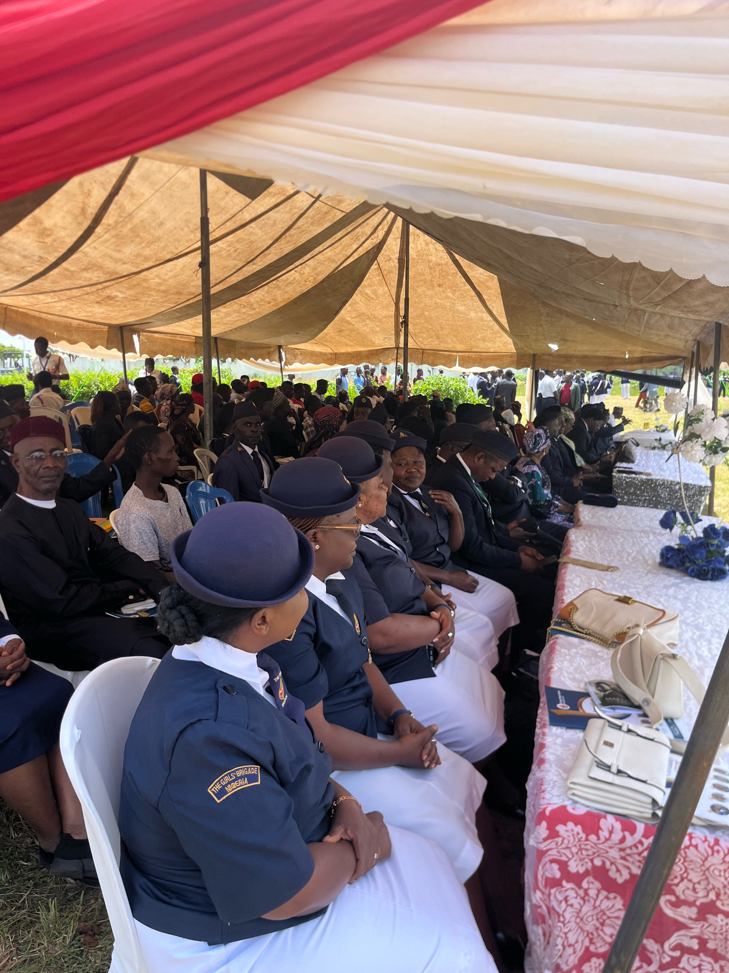 The image shows an outdoor gathering under a tent, likely a solemn occasion, given the presence of what appear to be coffins on a table in the foreground. A group of people are seated in chairs, facing a central point. Many of the individuals, particularly in the foreground and middle ground, are wearing uniforms. Several women in the foreground are wearing navy blue uniforms with white collars and hats, and one has a patch on her sleeve that reads "THE GIRLS BRIGADE NIGERIA". Men in the foreground are wearing dark suits and caps or hats. The tent has a red and white canopy overhead, and the environment appears to be grassy. The overall atmosphere suggests a formal event, possibly a funeral or memorial service, taking place in Barikin Jirga Summa, Nigeria.