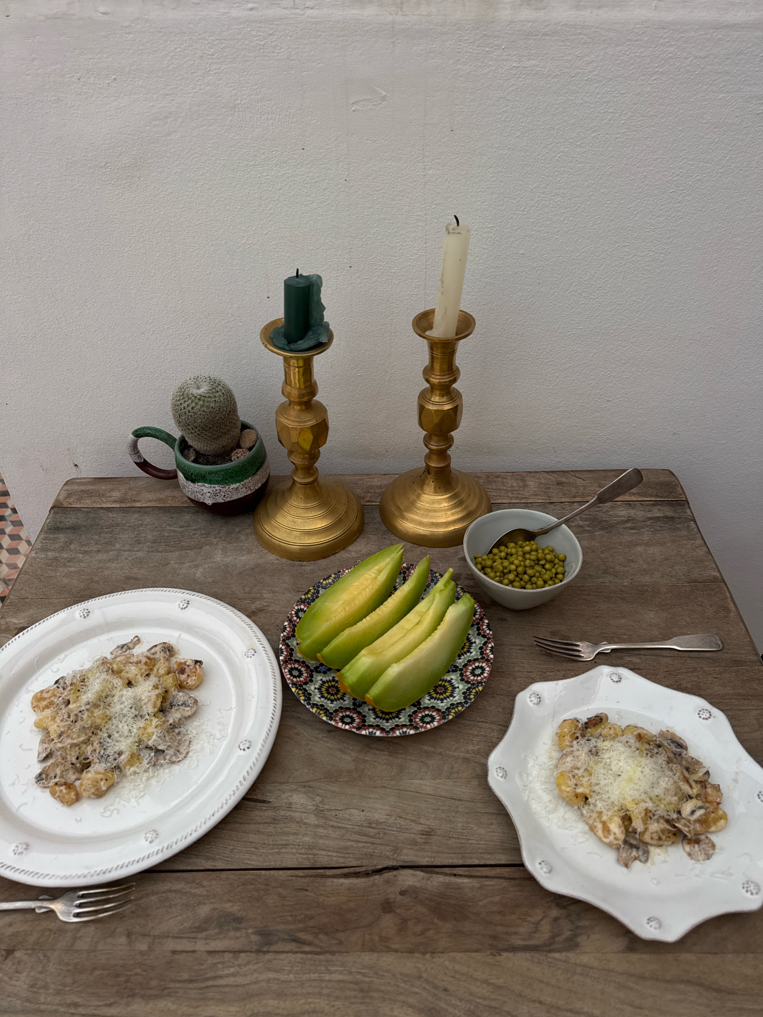 This image depicts an outdoor dining setting on a rustic wooden table against a white textured wall. Two plates of pasta, topped with grated cheese and mushrooms, are prominently featured. To the right of the pasta, a small bowl contains green peas. In the center, a decorative plate holds slices of a light green melon. Two brass candlesticks, one holding a partially burned green candle and the other a partially burned white candle, stand behind the food items. A small potted cactus, situated in a decorative ceramic pot, is positioned to the left of the candlesticks. A fork rests on the table near the right plate of pasta, and another fork is visible on the left plate. The overall scene suggests a meal prepared for two, set in what appears to be a serene outdoor location. The mention of Essaouira, Morocco, suggests this could be a scene from a Riad or a similar traditional Moroccan dwelling with an open courtyard or patio area.