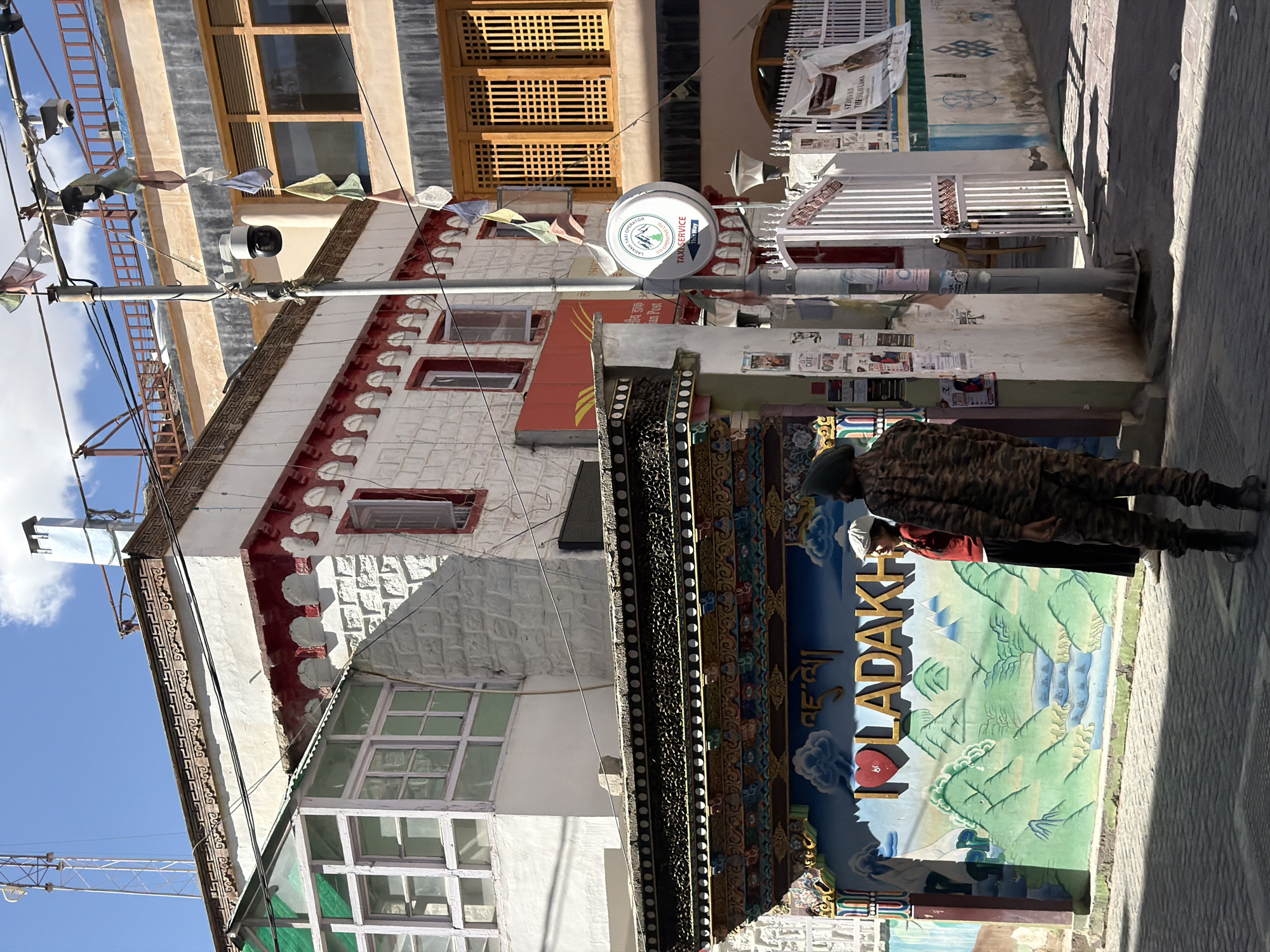 The image depicts a street scene in the city of Leh, India. In the foreground, two individuals are visible. One person, dressed in a camouflage uniform with a dark green cap, stands facing away from the camera. The second individual, wearing a red top and a white cap, is positioned to the left of the uniformed person and is looking towards the right.
Behind them, a vibrant mural adorns a wall, featuring green mountains, a river, and the text "I LOVE LADAKH" in large golden letters with a red heart symbol replacing the letter "O." The mural is framed by ornate, colorful architectural details.
The background reveals multi-story buildings with distinctive architecture. One building has white brick walls and red decorative trim. Another building features large wooden-framed windows. Prayer flags are strung across wires above the street, and a security camera is mounted on a pole. A white gate with vertical bars is visible on the right side of the image, leading to an entrance. Various posters and signs are affixed to walls and poles. The ground is paved and shows shadows cast by the buildings, indicating a sunny day. The overall environment suggests a busy, culturally rich urban setting.