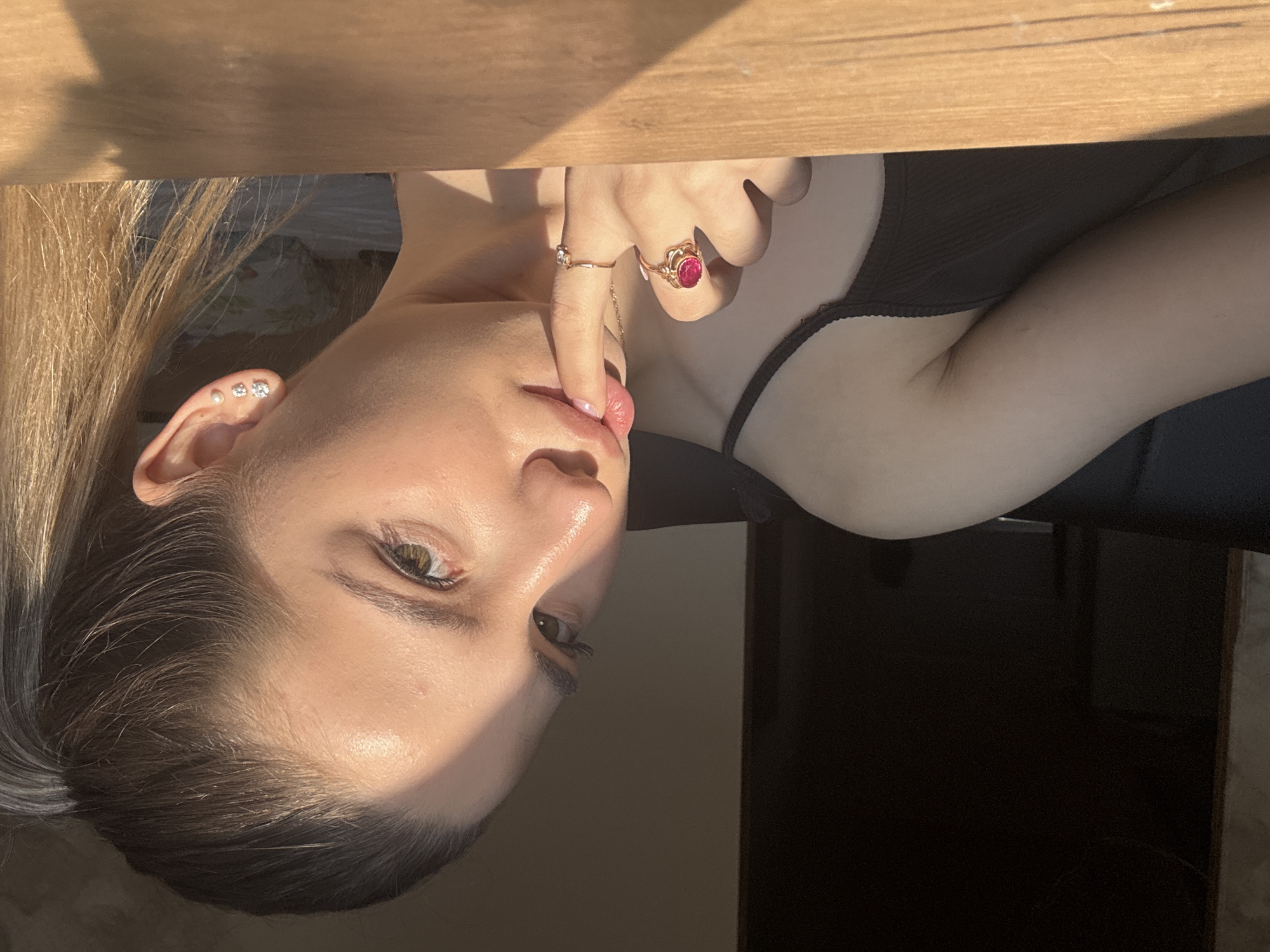A woman with light brown hair is positioned with her head tilted downward and facing toward the viewer. She has fair skin and green eyes. Her right hand is raised with her index finger touching her lower lip. She is wearing two rings on that hand, one with a small, delicate design and the other with a larger red stone. She is also wearing multiple earrings in her left ear. She has on a black top. A wooden surface is seen above her, suggesting she may be under a table. The background appears to be a dimly lit room with a dark piece of furniture. The location in the city of Noviy, Russia is not discernible from the given image.