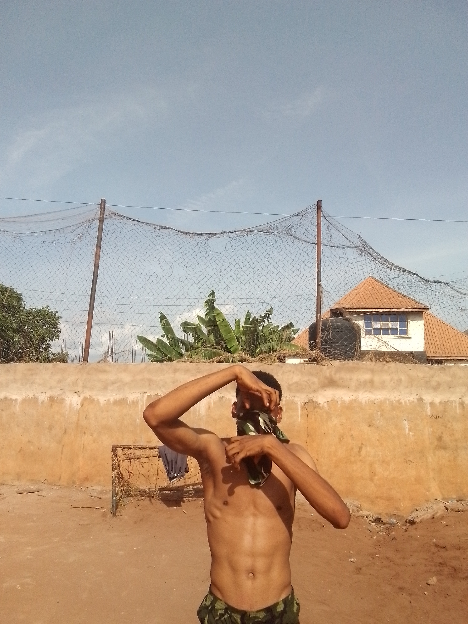 A shirtless man with dark skin and a camouflage bandana around his face is standing in a dirt yard. He has dark hair and appears to be thin. The yard is surrounded by a low, tan-colored wall. Behind the wall is a wire fence supported by metal poles. Beyond the fence are banana trees and a building with a dark water tank and a terracotta-colored roof. The sky is light blue.