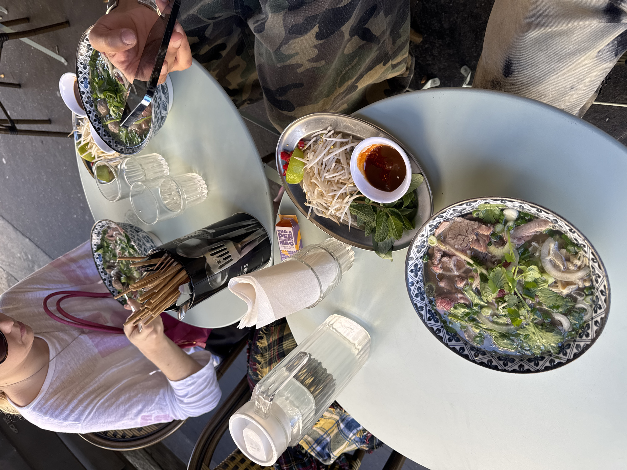 This image captures an overhead view of two people dining outdoors at a cafe in Paris, France. Two light-colored round tables are laden with bowls of Vietnamese pho, accompanying condiments, and drinks, suggesting a casual meal.
In the upper left, one person, wearing camouflage pants, is using metal tongs to handle ingredients within a bowl of pho, which contains meat, noodles, and fresh green herbs. Adjacent to this bowl are several empty clear glasses and a lime wedge.
On the table to the right, a prominently displayed bowl of pho is visible, featuring slices of meat, onions, cilantro, and broth. Alongside it on a small metal plate are fresh bean sprouts, lime wedges, sliced chili peppers, and mint leaves, accompanied by a small white bowl of dark red chili sauce.
In the lower left, another diner, wearing a white long-sleeved top and sunglasses, holds chopsticks, poised to eat. A red or pink purse is visible near their arm. Centrally located on the table is a black cylindrical utensil holder filled with wooden chopsticks and metal cutlery. A white napkin dispenser and a clear plastic pitcher filled with water are also present. A plaid blanket is draped over what appears to be a chair or lap.
A small, orange and purple product box is partially visible, displaying the text "THC-A PEN MAC" and "MIRACLE ALIEN". The bright lighting suggests it is daytime, creating a relaxed and pleasant atmosphere suitable for outdoor dining.