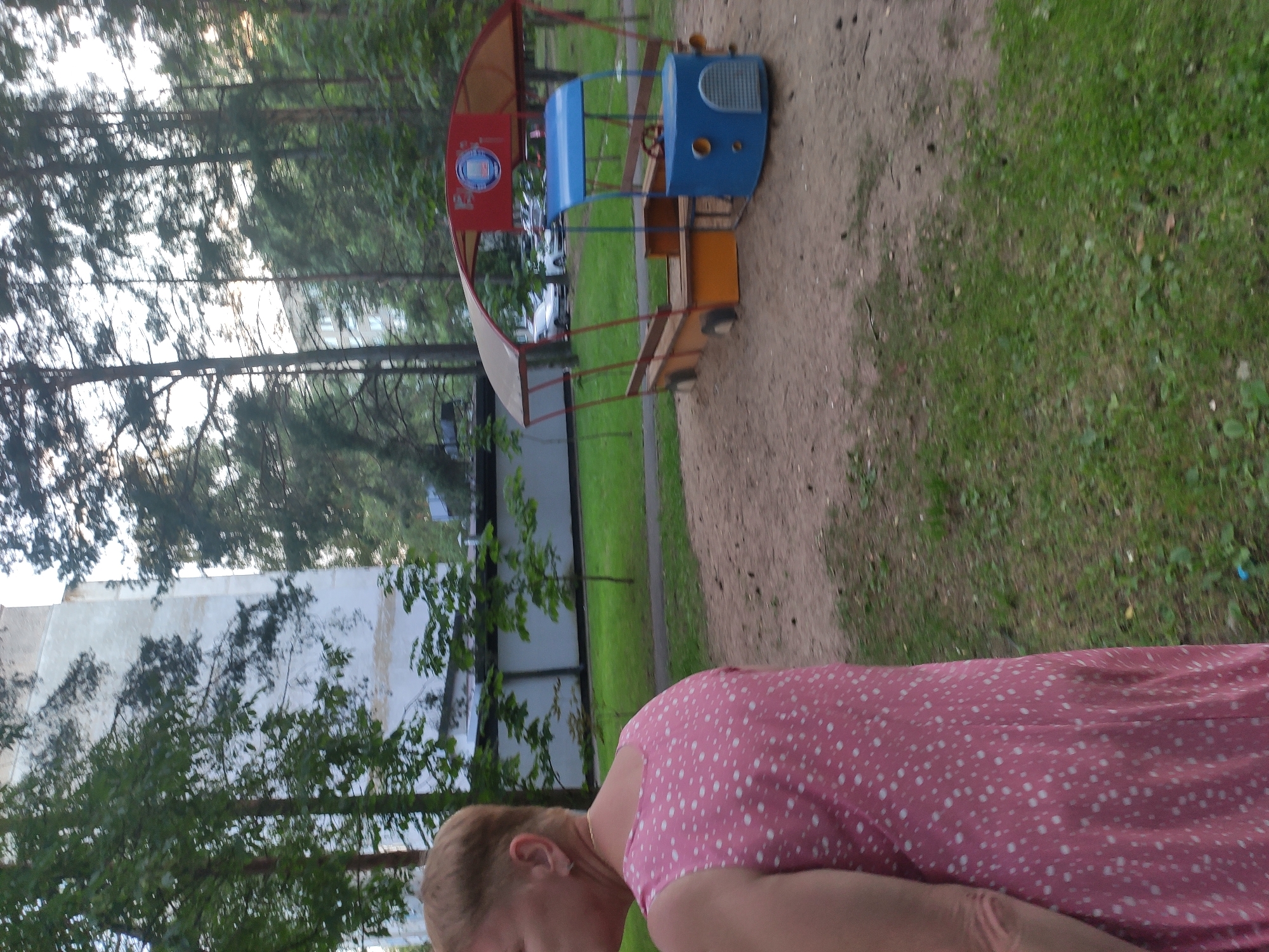 In the city of Navapolatsk, Belarus, a woman with short blond hair, wearing a pink dress with white polka dots, stands on the left side of the frame. She is partially visible from the shoulders up, with her back to the camera. In the background, a sandpit playground with a toy bus structure is visible. The playground features a red and yellow awning, a blue front section resembling a bus with a steering wheel, and wooden benches. The ground of the playground is covered in sand. Beyond the playground, there is a grassy area with trees and a tall apartment building. Several cars are parked in the distance. The environment suggests a residential area or park.