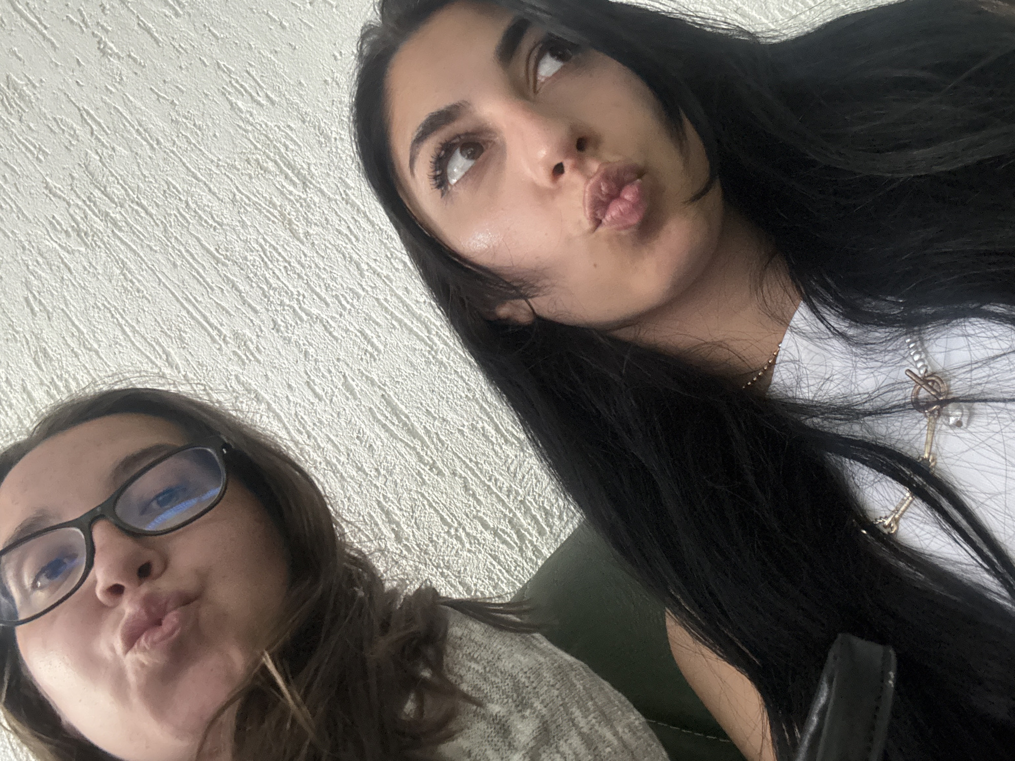 Two young women are positioned close to the camera, appearing to take a selfie. The woman on the right has long, dark hair, and she is looking up and to the left with her lips pursed. She is wearing a white top with a gold necklace. The woman on the left is wearing glasses and a gray sweater, and she also has her lips pursed. The background is a white wall with a textured surface. Given the lack of contextual information, it is impossible to ascertain if the image was taken in Plovdiv, Bulgaria. No further deductions can be made based on this limited visual information.