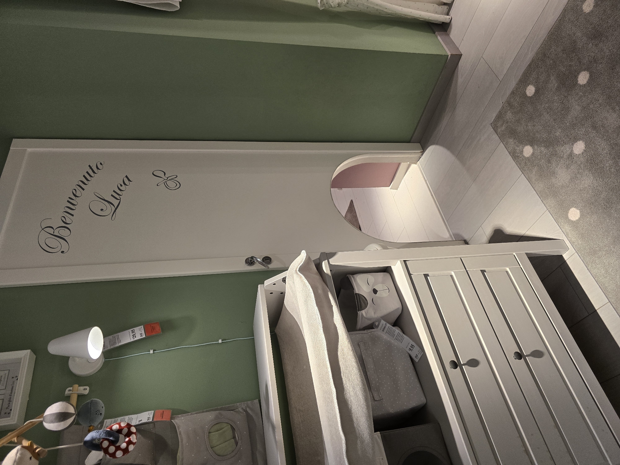 The image depicts a nursery room. A white changing table with drawers is on the left side of the frame. Above the changing table, a white wall lamp is mounted, with a price tag hanging from it. A baby mobile with stuffed toys is attached to the wall above the lamp. To the right, a white door is partially visible, with decorative cursive lettering that reads "Benvenuto Luca" and a pacifier illustration. The door has an arched opening at the bottom, revealing a pink-colored space beyond, with a light-colored wooden floor. The walls are painted in a muted green. A grey rug with white polka dots is on the floor in the foreground. The scene is indoors and appears to be a staged display, possibly in a furniture store or showroom in Baronissi, Italy. There are no people or animals present in the image.