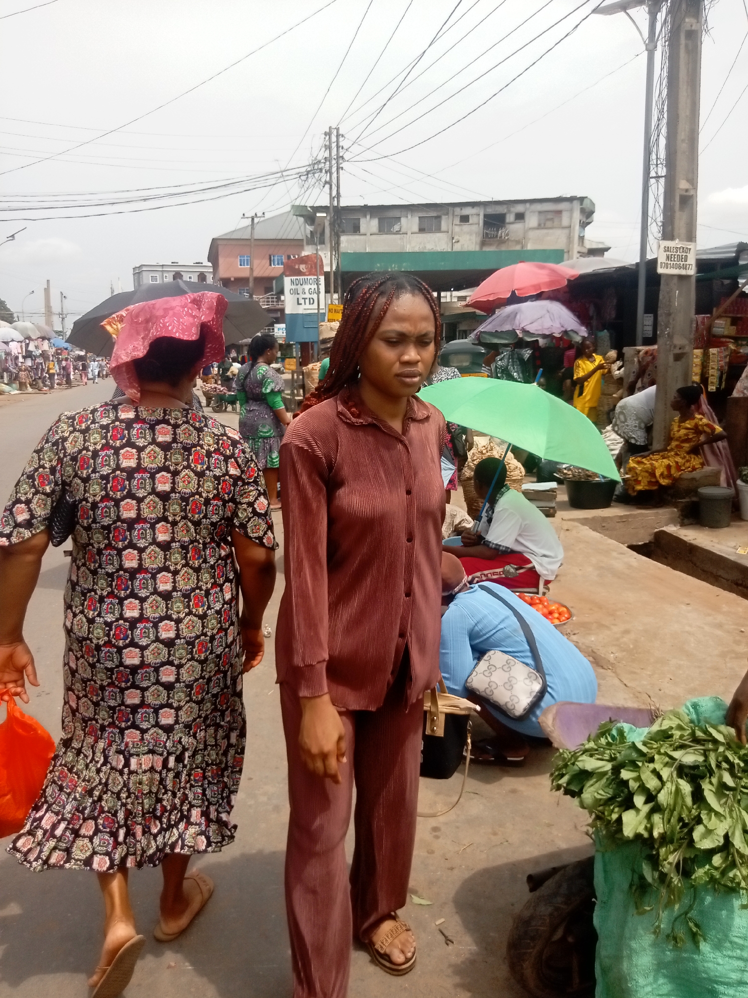The image depicts a bustling street scene in Onitsha, Nigeria, marked by commercial activity and the presence of numerous people. A woman in a brown, ribbed shirt and trousers stands in the foreground, her expression suggesting contemplation. To her left, another woman is seen walking away, carrying an orange bag and wearing a colorful patterned dress. The backdrop of the scene reveals various vendors situated along the street, many sheltered under umbrellas of various colors. They sit or kneel beside their wares, offering items such as produce for sale. The environment is characteristic of an urban market setting, with power lines overhead and buildings and signage visible in the distance, indicating the city location. Vegetation is also visible, hinting at locally grown produce being sold in the market.