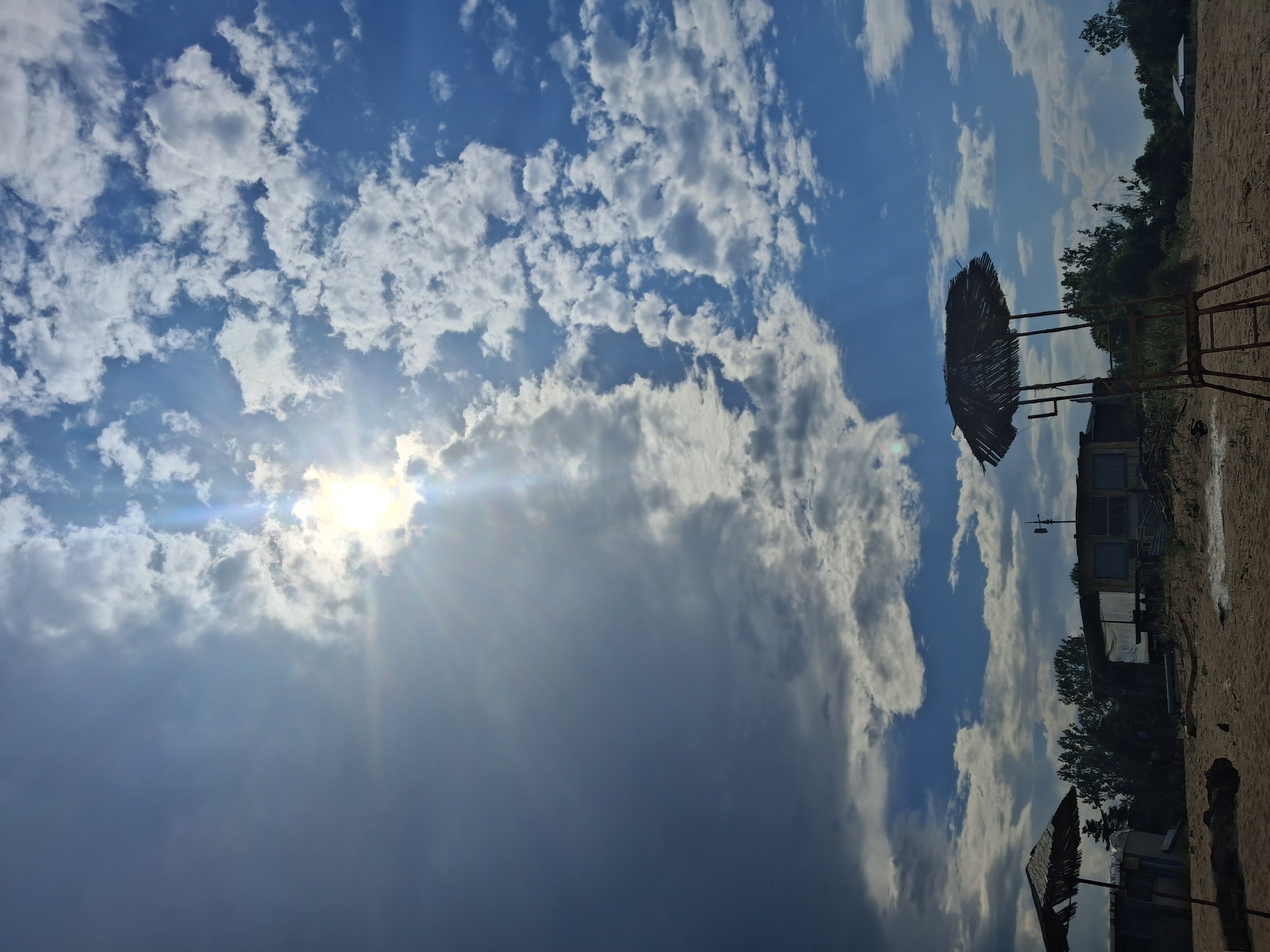 This image captures a beach scene in Shkorpilovtsi, Bulgaria, under a partially cloudy sky. The sun is visible in the upper center-right, partially obscured by clouds, emitting strong rays. The sky transitions from a darker, more overcast appearance on the left to a brighter blue with scattered white cumulus clouds on the right. The foreground consists of sandy terrain, featuring a prominent, worn thatched beach umbrella on a metal frame, possibly a lifeguard chair, positioned in the lower right-center. A second, partially visible and damaged thatched umbrella is situated on the far left. Behind the sandy area and a line of trees, several rustic or makeshift structures are discernible, including a building with what appears to be a wind vane or antenna on its roof. No individuals are present in the scene.