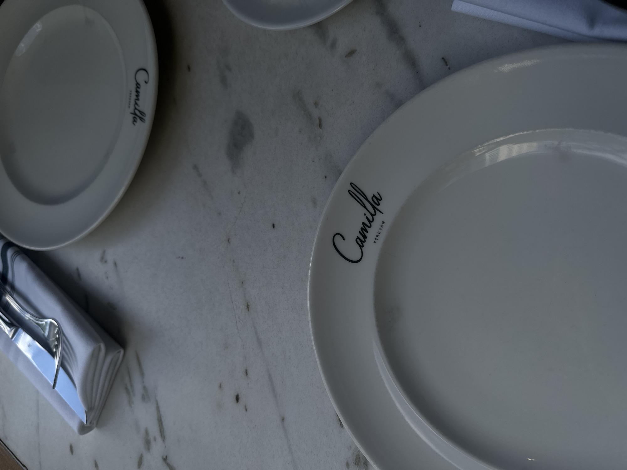 The image shows a restaurant table setting with white marble and plates branded with "Camilla Yerevan", indicating the location is in Yerevan, Armenia. There are two visible plates with the "Camilla Yerevan" logo and cutlery wrapped in a white napkin on the table.