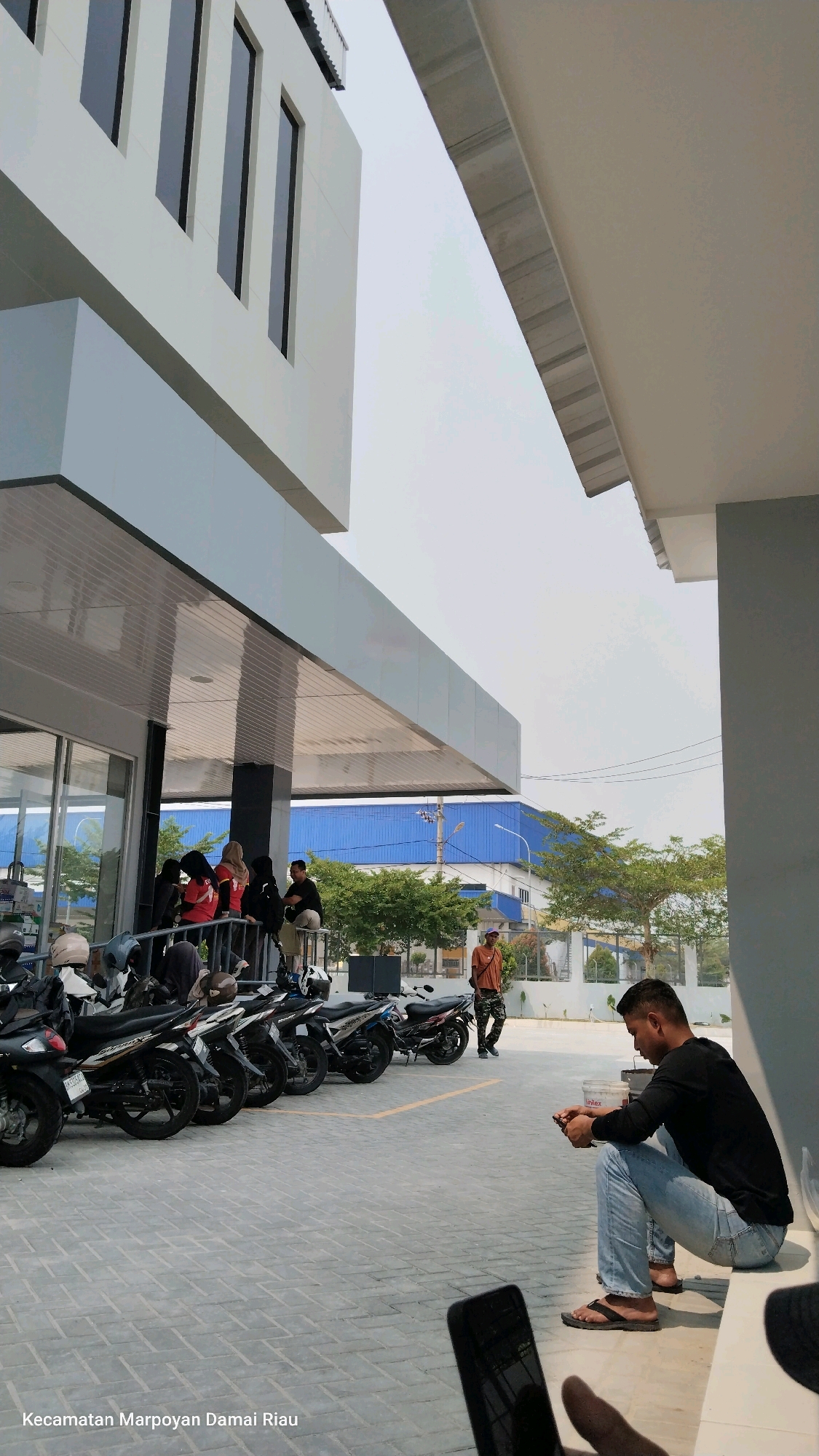 The image shows a group of people and motorcycles outside a modern building, possibly a business or office. The building has a white facade with dark-framed windows on the upper floor and a cantilevered roof structure with a metallic ceiling. A man in a black long-sleeved shirt and blue jeans is sitting on a ledge, looking at his phone. A few people, some wearing red shirts and headscarves, are standing under the roofed area. A row of parked motorcycles is visible in the foreground. In the background, there are trees and a blue industrial building. The scene is set on a paved area, likely a parking lot. The text at the bottom of the image, "Kecamatan Marpoyan Damai Riau," indicates the location is Marpoyan Damai District in Riau, Indonesia, which is part of the city of Pekanbaru.