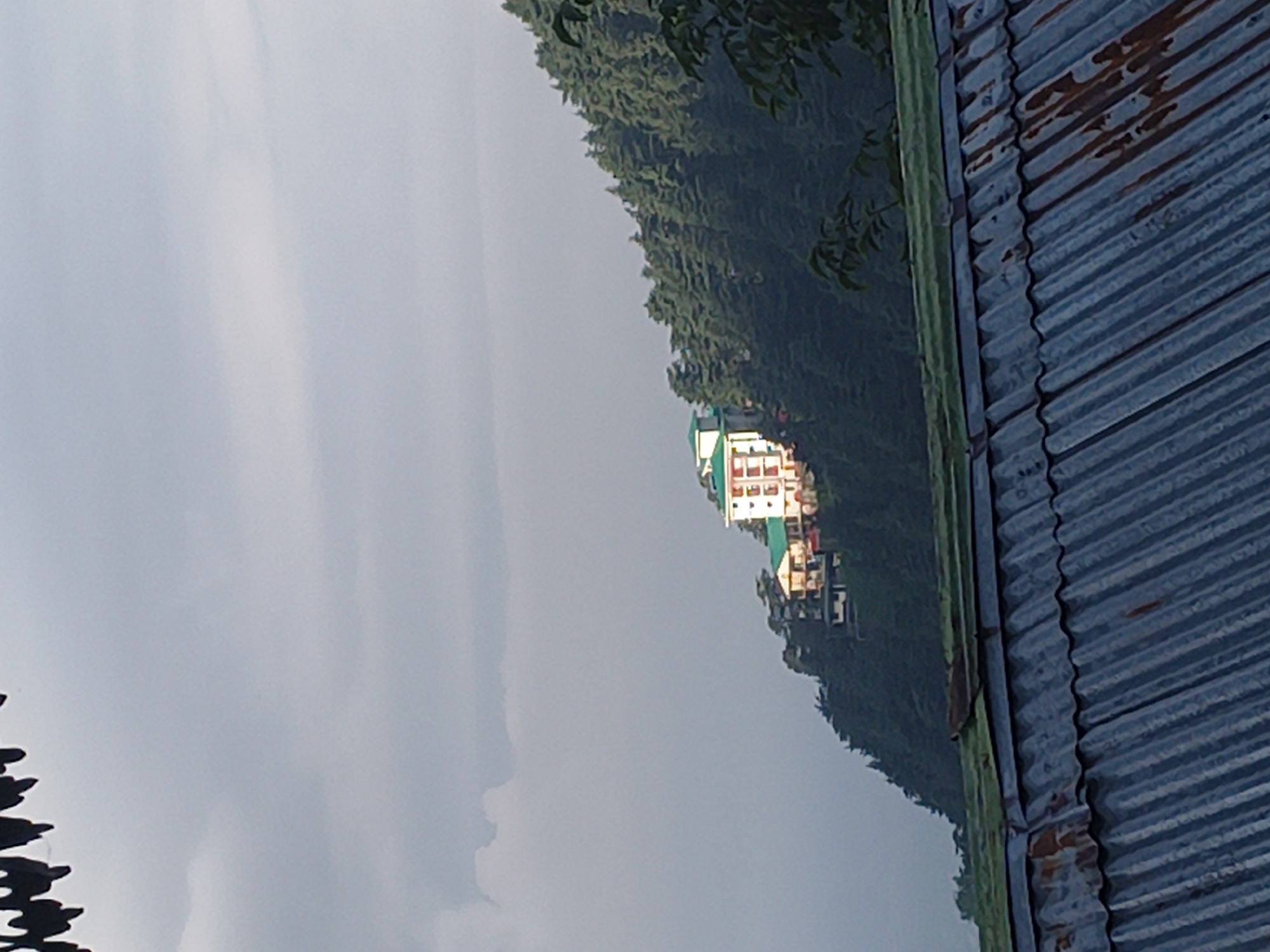The image shows a multi-story building with a green roof, perched on a lush, green hillside covered in coniferous trees. The building appears to be illuminated, possibly by the sun breaking through overcast skies. In the foreground, a corrugated metal roof with patches of rust is visible, suggesting it might be part of a structure in the foreground that is closer to the viewer. The overall scene depicts a mountainous or hilly terrain, likely in a rural or semi-rural area, with the building standing out against the natural landscape. The lighting conditions suggest it may be early morning or late afternoon.