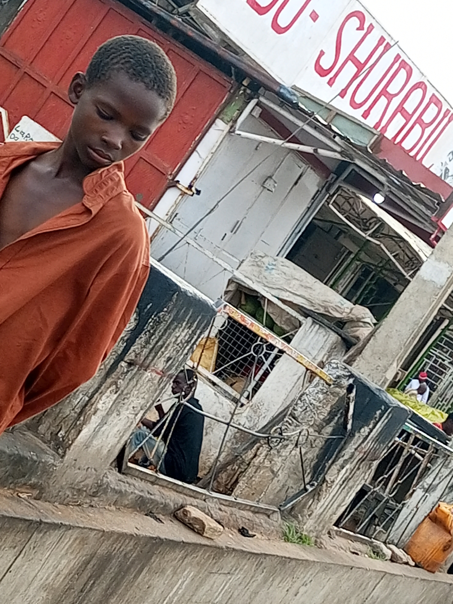 A young boy, appearing to be of African descent, stands in the foreground, looking down with his head tilted. He wears an orange, open-collared shirt. The background depicts a marketplace or a row of shops. A large red sign with white and red lettering, partially obscured, is visible above the shops. The shops appear to be constructed of corrugated metal and wood, with some areas covered by tarps. In the middle ground, behind a metal railing, a person in a dark garment is seated. Further in the background, other individuals are faintly visible among the stalls. The overall scene suggests an outdoor urban environment, possibly a bustling market.