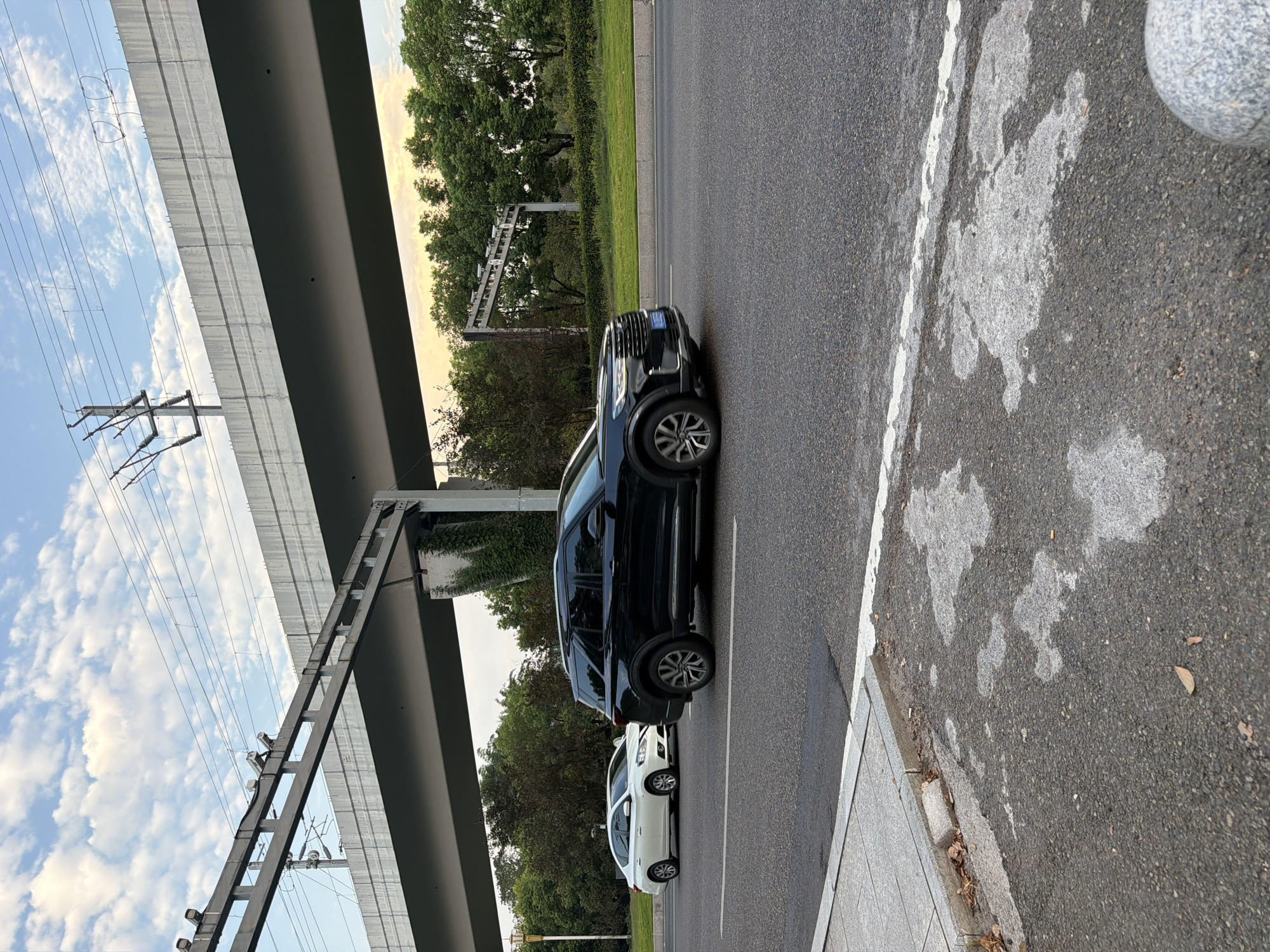 In Jiangjin, China, a black SUV is traveling on a multi-lane road beneath a concrete overpass. A white sedan is also on the road, slightly ahead of the SUV. The overpass has overhead wires and what appear to be surveillance cameras mounted on it. The scene is framed by green trees and a sky with scattered clouds. The foreground shows the asphalt road surface with white lane markings and a sidewalk made of concrete tiles.