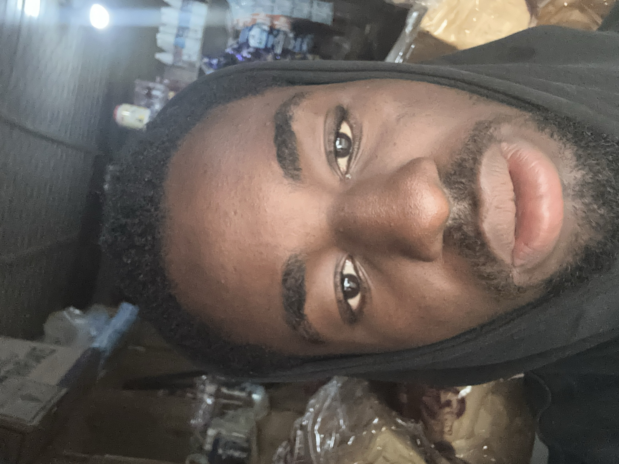 A close-up, head-on selfie of a Black man wearing a black hooded garment. The man's face fills most of the frame, with his eyes looking directly at the camera. His skin is dark brown, and he has short, dark, curly hair. He has a light beard and mustache. The background is out of focus, appearing to be an indoor setting with shelves stocked with various items, possibly in a store or market. The lighting is bright, with a light source visible in the upper right quadrant of the image.