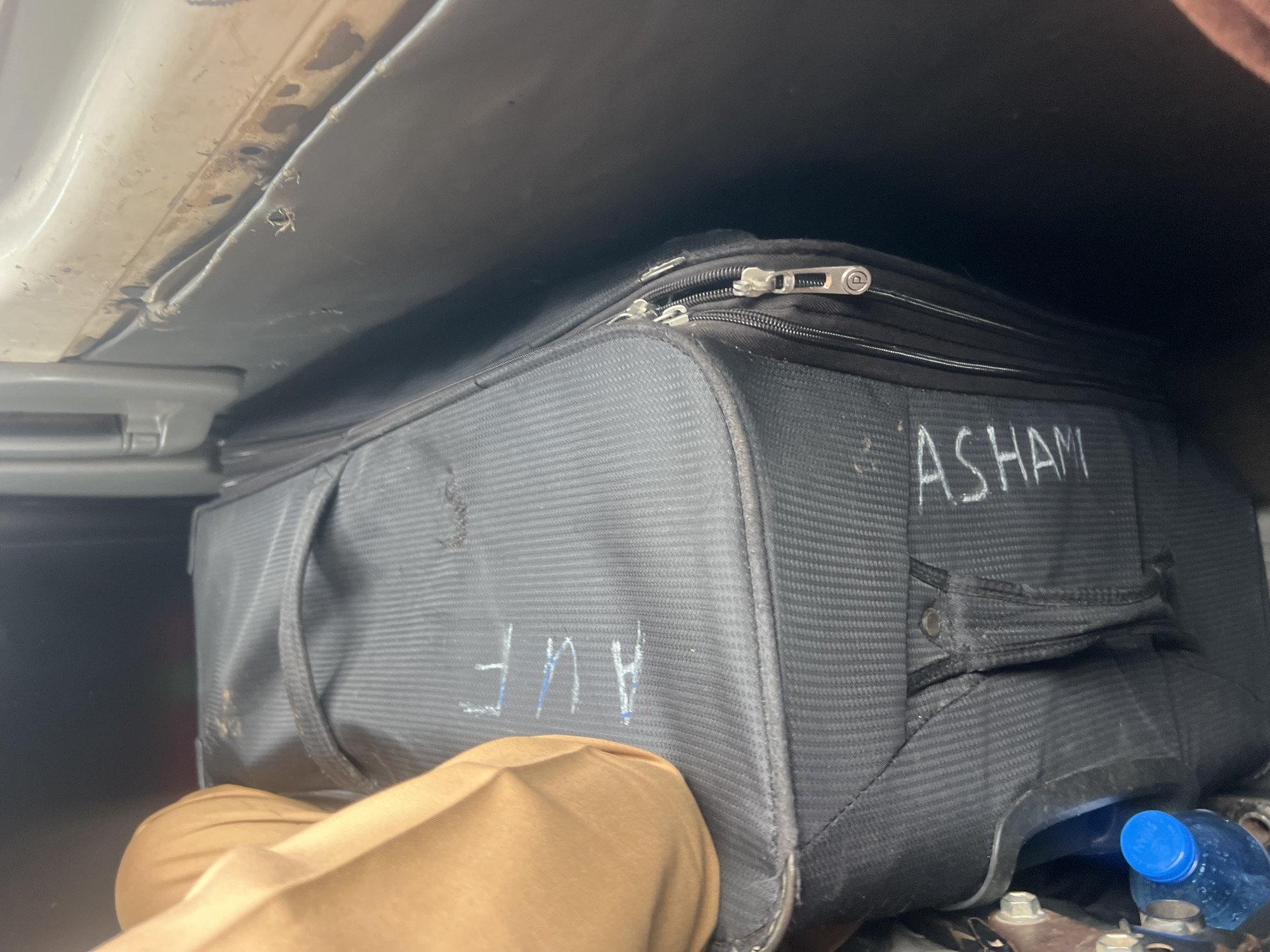 A black suitcase with the names "AUF" and "ASHAM" written on it in white marker sits in the back of a vehicle. A tan fabric is draped over the left side of the suitcase. A blue-capped water bottle is visible at the bottom left of the image. The scene appears to be in Makera, Nigeria.
