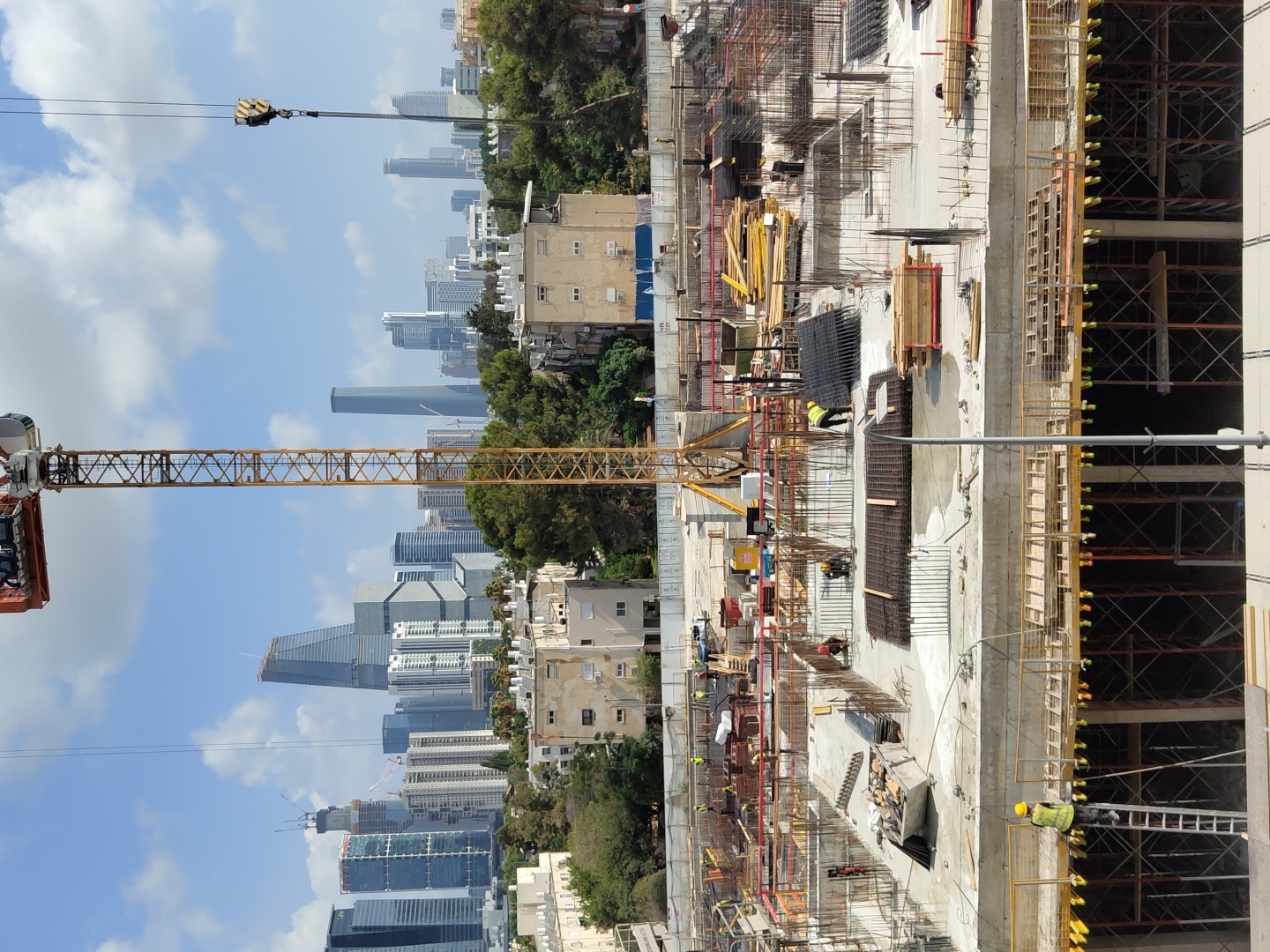 The image depicts an ongoing construction site in Tel Aviv, Israel, with a cityscape in the background. A prominent yellow construction crane dominates the center of the frame, with its boom extending upwards. The foreground of the construction site is filled with scaffolding, rebar, concrete, and construction materials. Several workers in high-visibility vests and hard hats are visible, engaged in various construction activities. One worker is ascending a ladder on the left side of the frame, while others are seen working on different levels of the site, some handling materials and others overseeing the work. The midground shows a building under construction, with exposed structural elements and concrete surfaces. In the background, the Tel Aviv skyline is visible, featuring a collection of modern skyscrapers and residential buildings, indicating an urban environment. The sky is partly cloudy, suggesting daytime. The overall scene conveys a sense of industrial activity and urban development.
