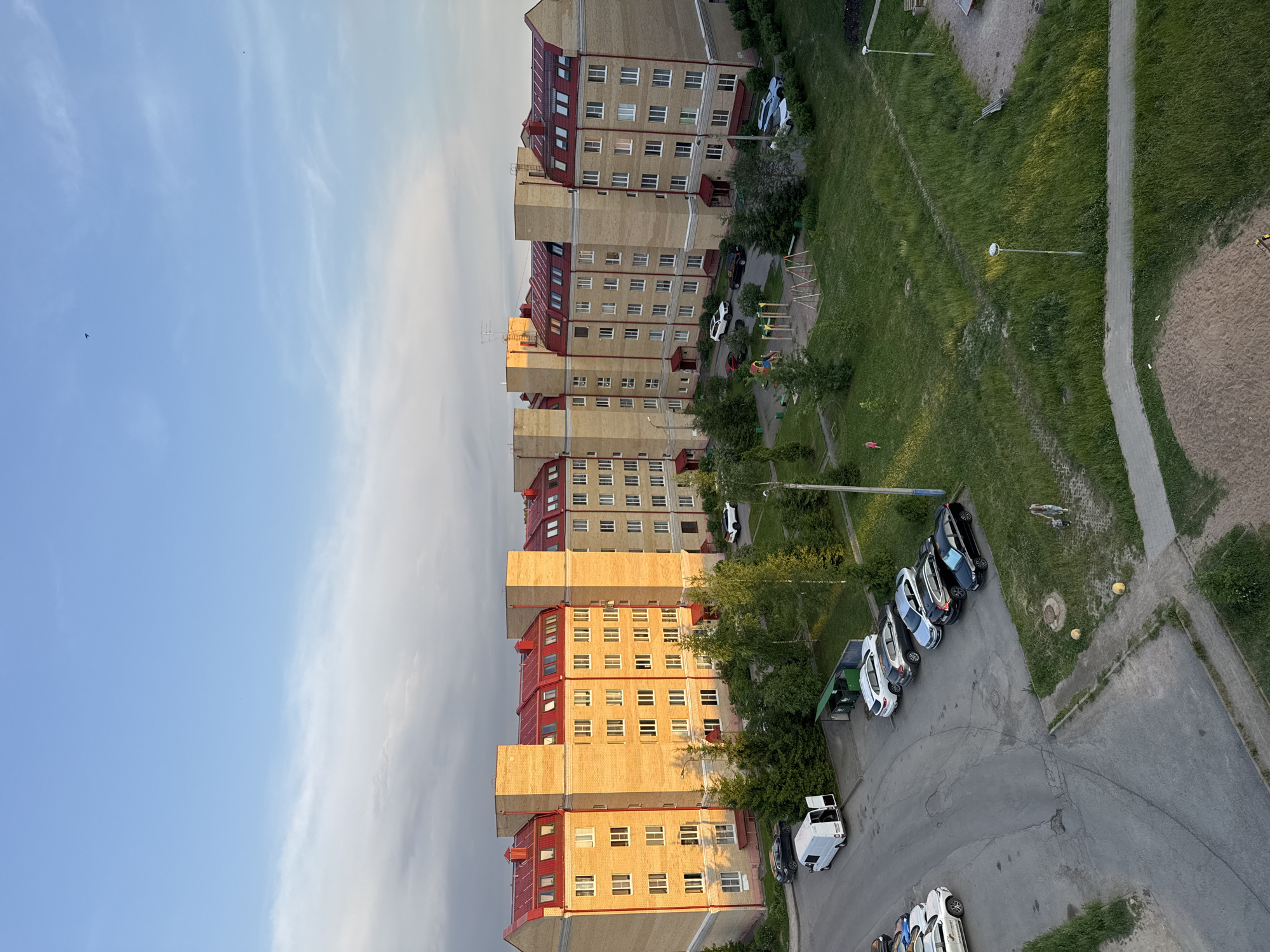 The image captures an aerial view of a residential area in Kirishi, Russia, likely taken during late afternoon or early evening, as indicated by the warm, golden light illuminating the buildings. The dominant feature is a long, multi-story apartment building constructed with light yellow bricks and a distinctive red-tiled, mansard-style roof. The building is segmented into several identical sections, each with multiple floors, numerous windows, and small dormer windows on the upper level.
In front of the building, a paved area serves as a parking lot where several cars are parked, including a white van with its rear doors open. Adjacent to the parking area is a strip of green grass, interspersed with trees and bushes. A children's playground with swings and colorful structures is visible within the green space. Further to the right, more grass and trees are present, along with streetlights and a pathway. The sky above is a clear blue with wispy white clouds, and a small bird is visible in flight. The overall scene depicts a typical urban residential environment with modern architecture and common amenities.