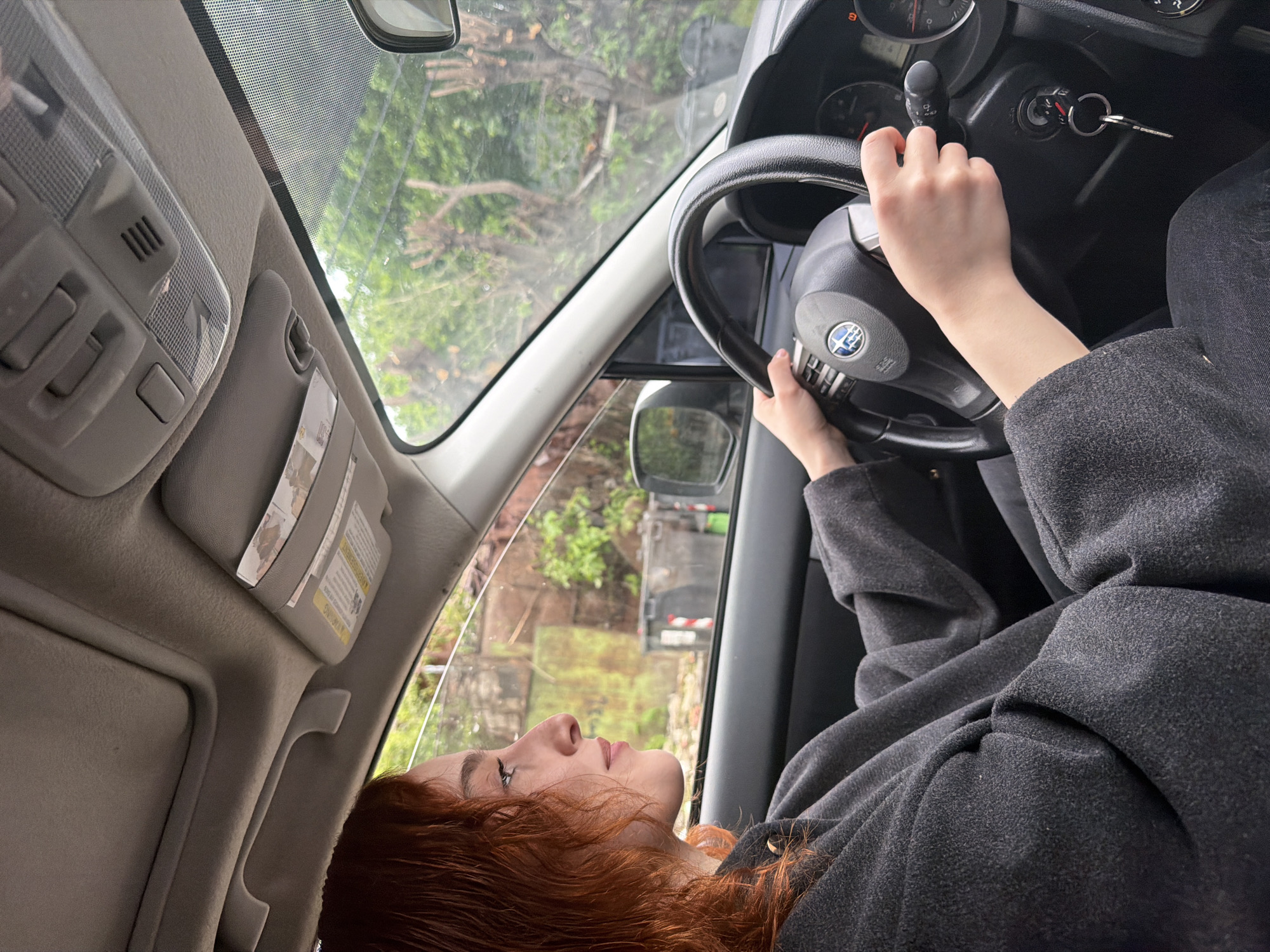 A woman with red hair is sitting in the driver's seat of a car. She is wearing a grey coat and has her hands on the steering wheel. The car keys are in the ignition. Through the windshield, trees and buildings can be seen. Given the style of buildings and general environment visible through the windshield, the location could be Tbilisi, Georgia.