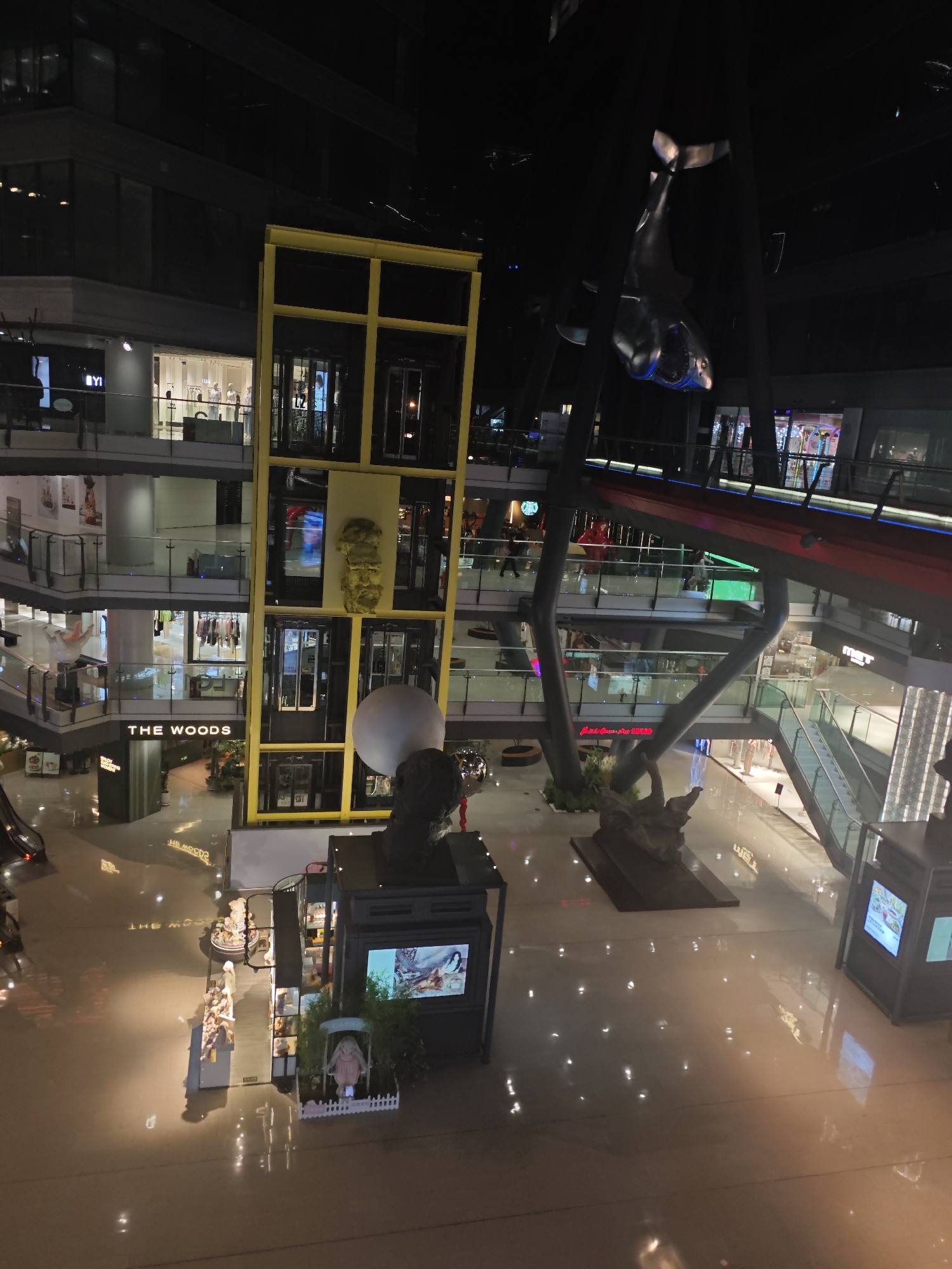 This image captures the interior of a modern, multi-story shopping mall or commercial complex at night or in the evening, located in Chaoyang District, China. The scene is illuminated by artificial lighting, which reflects on the glossy floor surfaces.
A prominent feature is a tall, bright yellow structural frame housing multiple glass-enclosed elevators, extending through several levels of the building. To the right, a large, dark sculpture of what appears to be a shark or similar marine creature hangs from the high ceiling. Below it, on a lower floor, another dark sculpture resembling a large animal, possibly a crocodile or alligator, is displayed. Near the base of the yellow structure, a large white sphere and a dark bust sculpture are visible.
The mall consists of multiple levels of shops, characterized by their illuminated interiors and display windows. Each level features glass railings along the walkways. Escalators are visible on the right side, connecting the various floors. A distinctive elevated walkway or bridge, featuring red and blue elements, spans across the upper levels of the complex.
On the ground floor, several displays and kiosks are present, including one with a white picket fence and a light-colored rabbit statue. A few distant figures of people can be seen within shops or on walkways, indicating general mall activity.
Visible text includes the shop name "THE WOODS" on a dark sign on one of the lower levels, and "YI" visible on another shop's sign. The architecture is modern and expansive, creating a spacious and contemporary atmosphere.