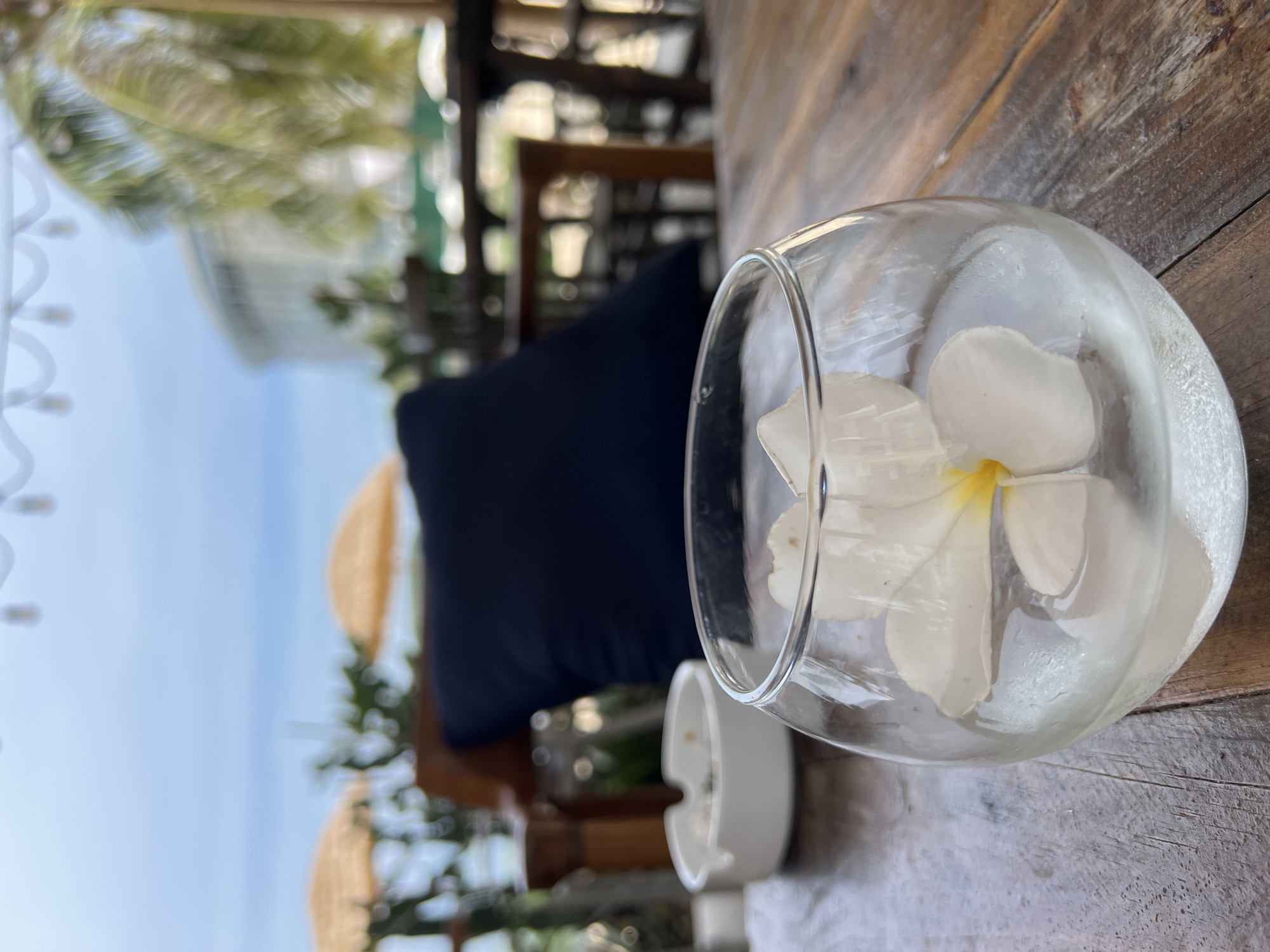 A close-up shot of a clear glass containing a white plumeria flower and ice cubes sits on a weathered wooden table. To the left, a white ceramic ashtray is visible. In the background, a dark blue cushion is placed on a wooden chair. Beyond the chair, two yellow umbrellas and palm trees are set against a hazy sky, suggesting an outdoor setting at a beach resort or cafe. The overall ambiance is relaxed and tropical. This scene is likely at a beachside establishment in Nha Trang, Vietnam, given the presence of palm trees and the clear sky often seen in coastal areas.