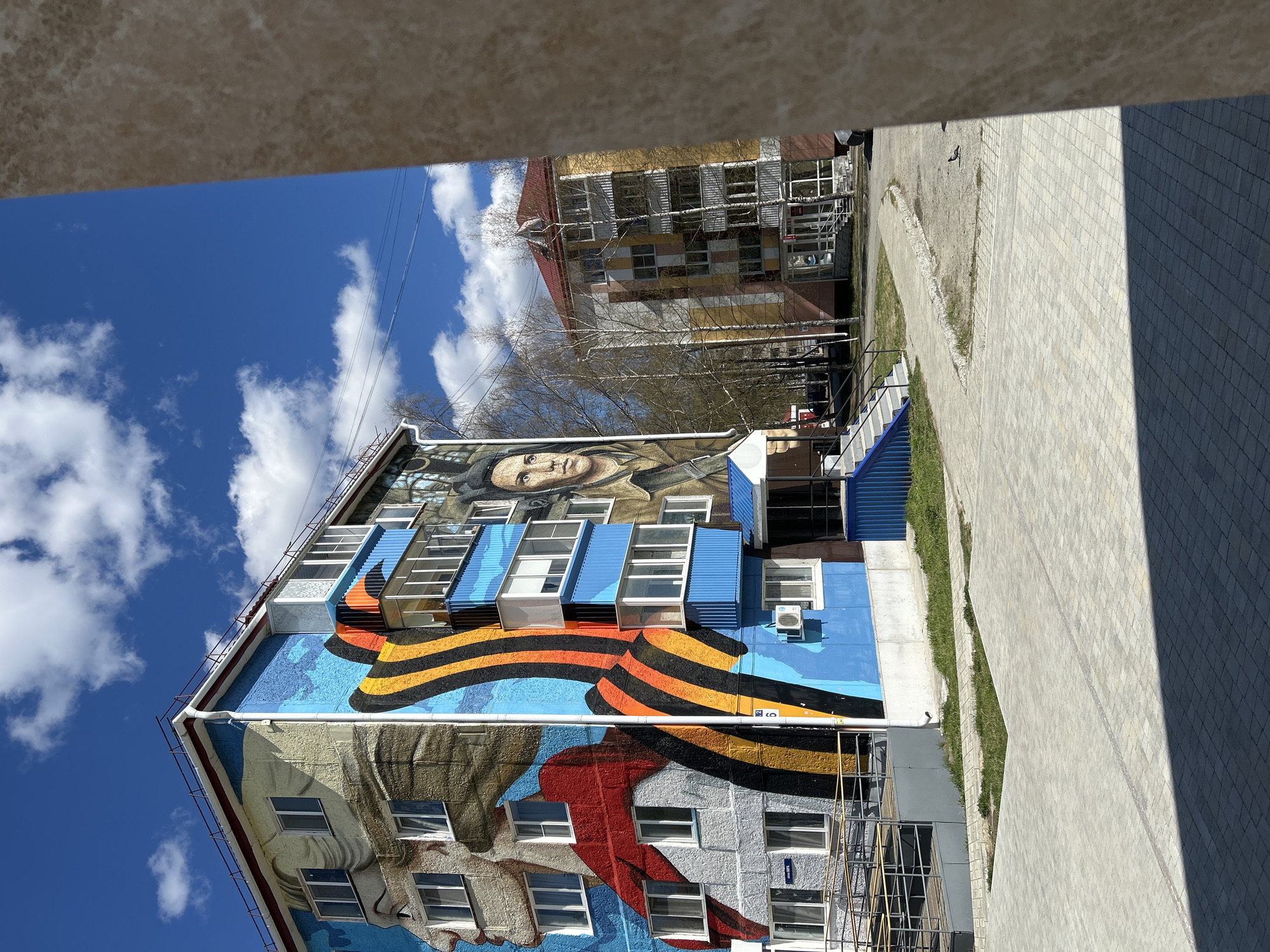 The image shows a building with a large mural depicting portraits and a St. George ribbon, located in Uray, Russia. The building has multiple windows, some with blue awnings. Scaffolding is present on the side of the building. In the background, another building with a multi-colored facade can be seen. The sky is blue with scattered clouds. Part of a balcony structure frames the top and right of the image. A paved walkway is adjacent to the building.