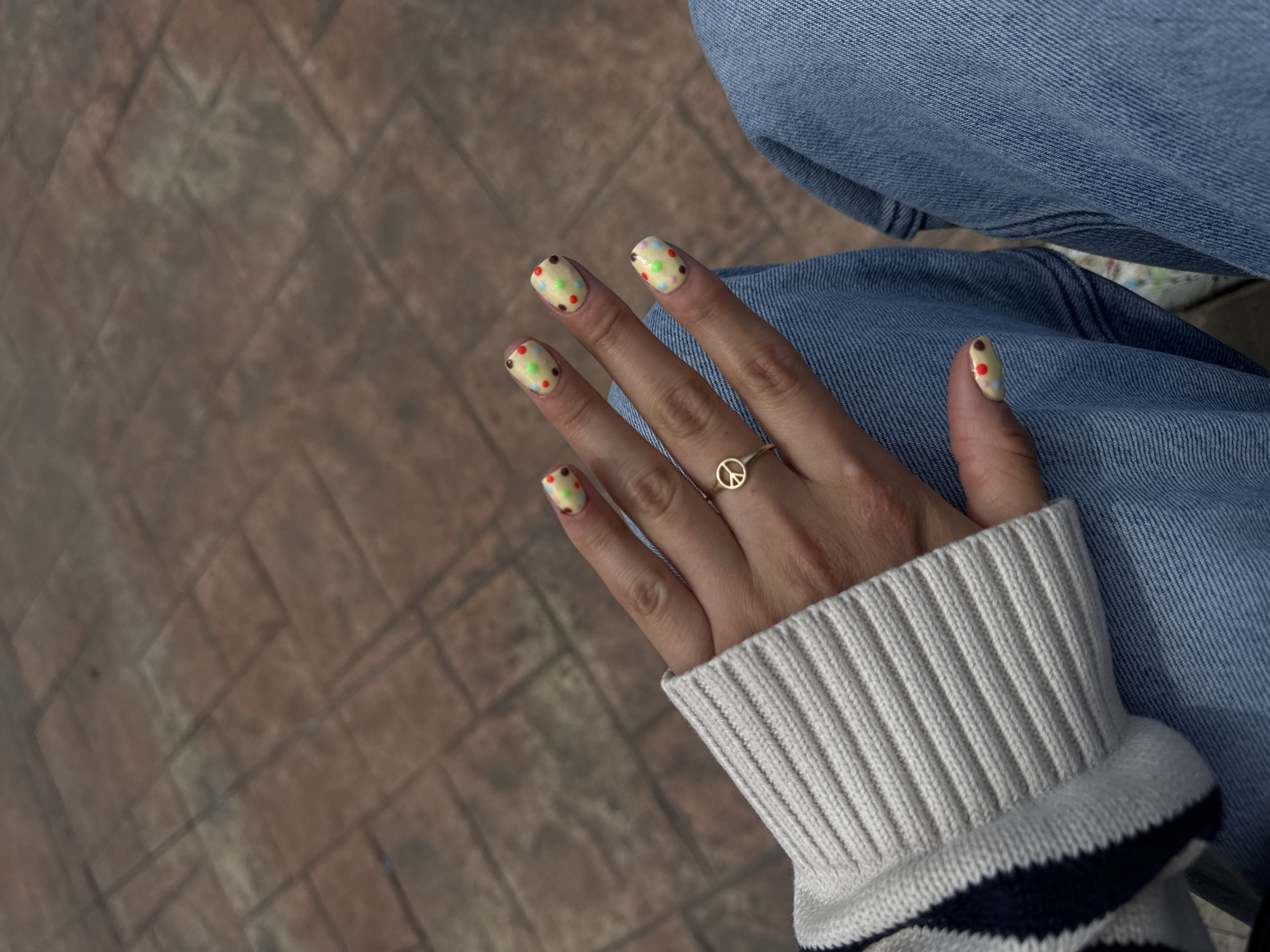 A high-angle shot shows a person's hand adorned with a gold ring bearing a peace symbol. The fingernails are painted with a pale yellow base and decorated with multicolored dots. The hand rests against denim-clad legs. The person is wearing a white sweater with navy stripes at the cuff. The background features a brick-paved ground. The location is implied to be in the city of Metepec, Mexico.