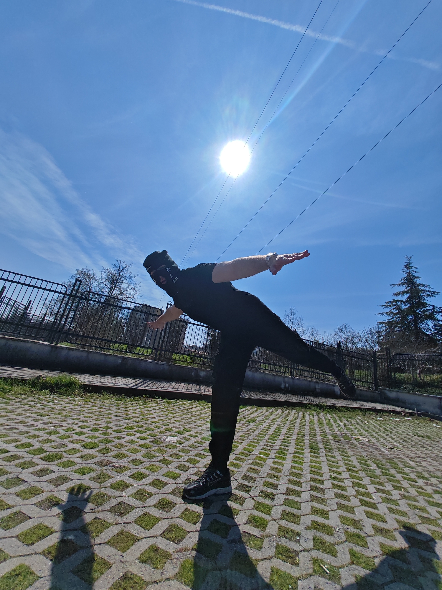 An individual is depicted performing a balancing pose outdoors in Shumen, Bulgaria. The subject is dressed in a black balaclava featuring white text, a black t-shirt, black trousers, and dark athletic shoes with light soles. A silver-toned watch is visible on the left wrist. The individual stands on their right leg, with the left leg extended straight backward and upward, and both arms extended horizontally outwards, resembling an airplane pose. The body is slightly tilted forward.
The scene is set on a surface paved with concrete grid pavers, allowing green grass to grow through the interstices. Distinct shadows of the individual are cast on this surface, indicating bright sunlight. A dark metal fence stretches horizontally across the midground, separating the paved area from a vegetated zone. Behind the fence, several leafless deciduous trees are present, alongside a prominent evergreen conifer on the far right. The sky is clear and blue, with faint contrails visible. A bright sun, exhibiting lens flare, is positioned in the upper right quadrant of the frame, illuminating the scene. Multiple dark, thin horizontal lines, consistent with power or communication lines, traverse the sky in the upper midground.