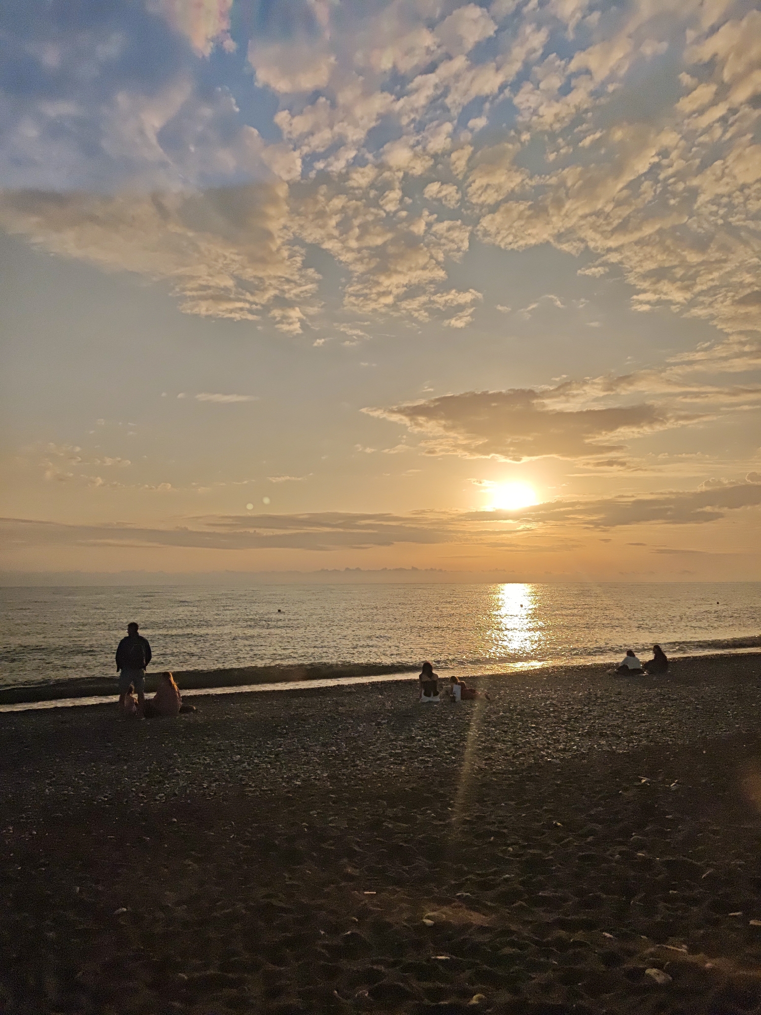 The image shows a beach at sunset in Sochi, Russia. The sky is filled with light clouds, with the sun setting on the horizon, creating a bright reflection on the water. Several people are on the beach, including a standing adult with two small children sitting down, another group of two adults with a child, and a separate group of two adults. The beach is made of dark pebbles and sand. The sea is calm with small waves.
