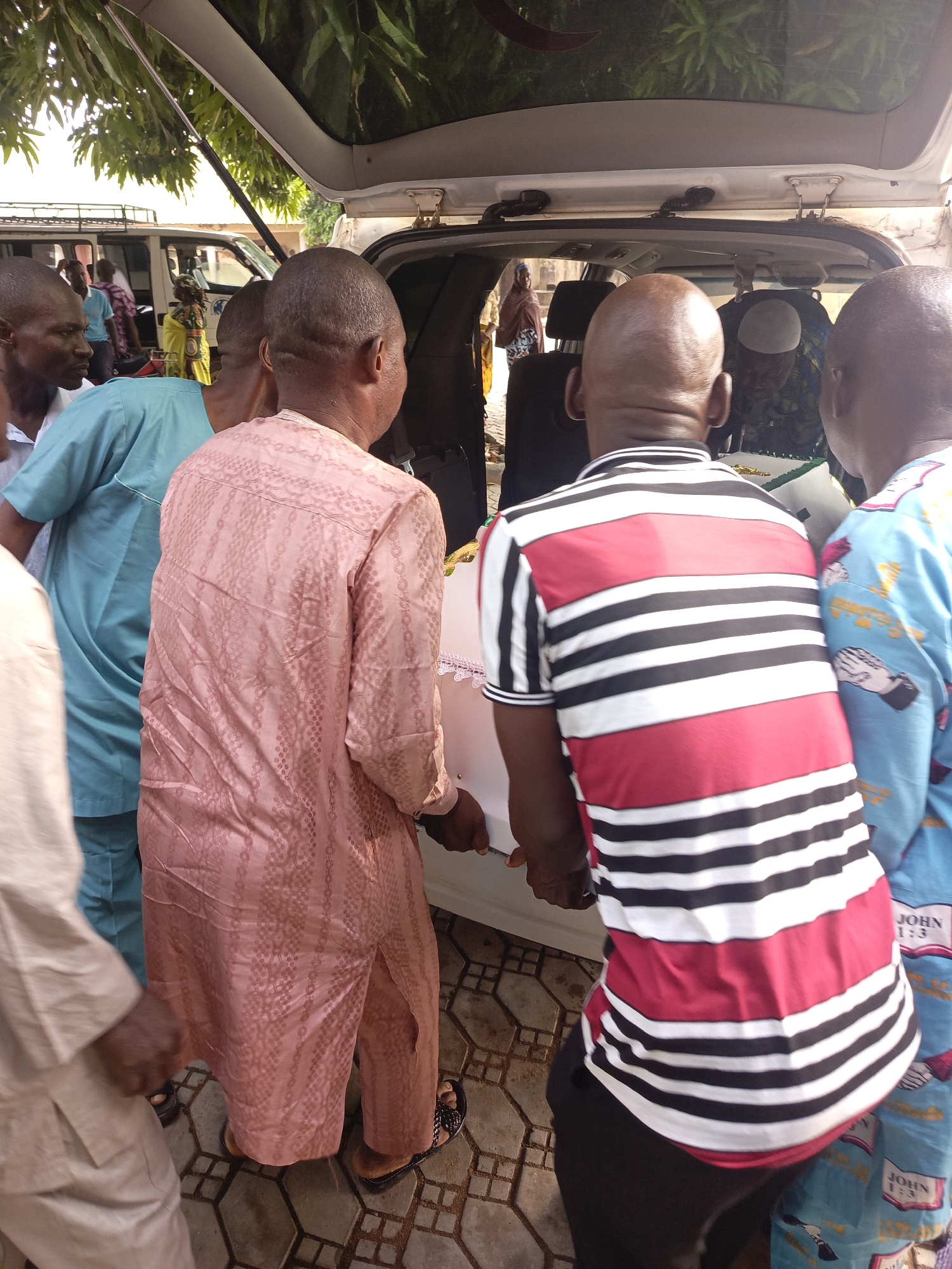 In a daytime scene in Akwanga, Nigeria, multiple men are engaged in the process of loading a white, rectangular object, likely a casket, into the open rear hatch of a light-colored vehicle, possibly a van or SUV. This activity suggests a funeral or burial event.
The foreground is dominated by the backs of several men. On the left, a man in a light-colored, possibly beige, traditional garment stands partially visible. Next to him, a man in a light blue tunic looks to his left. A prominent figure in the center wears a long-sleeved pink traditional garment with textured patterns. To his right, another man, seen from behind, wears a red, black, and white striped short-sleeved shirt. On the far right, a man in a patterned light blue garment, featuring repeated images and text, is also assisting. The text "JOHN 1:3" is clearly visible multiple times on his sleeve.
The white object being moved appears to be covered with a fabric or material, showing some decorative elements. The vehicle's interior is partially visible, with a dark headrest. Overhead, lush green foliage provides shade, indicating an outdoor setting. The ground is paved with hexagonal and square-patterned tiles.
In the background, other individuals, including a woman in a yellow dress and another in a brown headscarf, are faintly visible, along with parts of other vehicles. The overall atmosphere appears somber, consistent with a funeral procession.