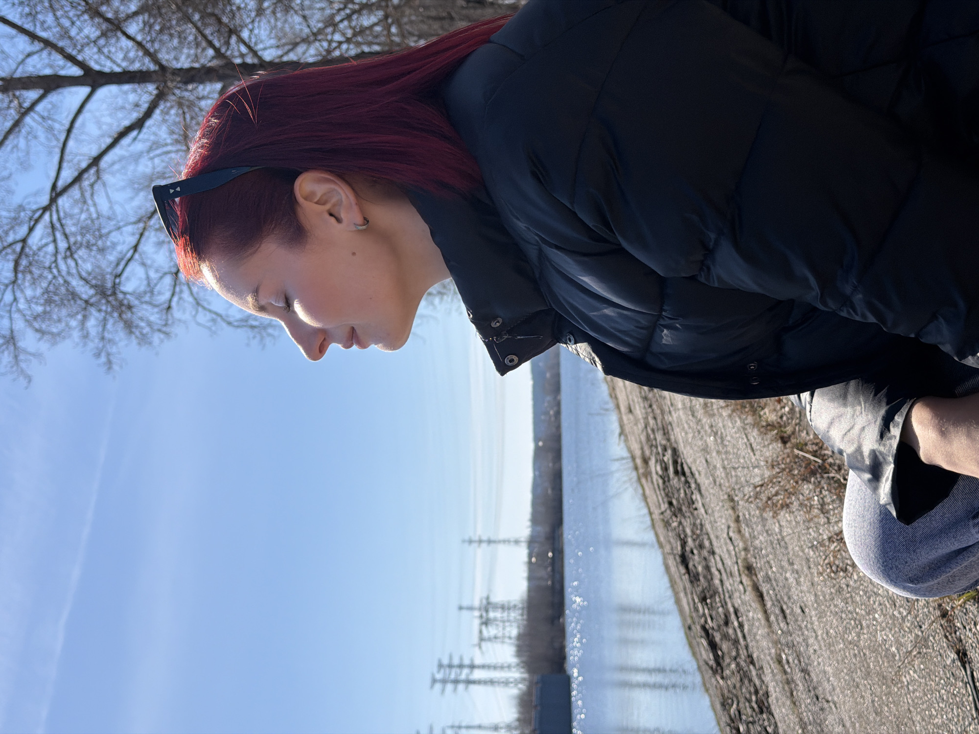 The image depicts a woman with auburn hair, wearing a black puffer jacket, gazing downwards. Sunglasses are perched on her head. She is standing near a body of water, possibly a river or lake, as evidenced by the distant reflection of power lines in the water. A concrete bank or embankment is visible to her right. The sky is clear and blue, suggesting a sunny day. The presence of power lines and a treeless winter landscape points towards an industrial area within a colder climate. The location is most likely in the city of Chaikovsky, Russia, given the prompt. The woman is interacting with the environment by observing the water.