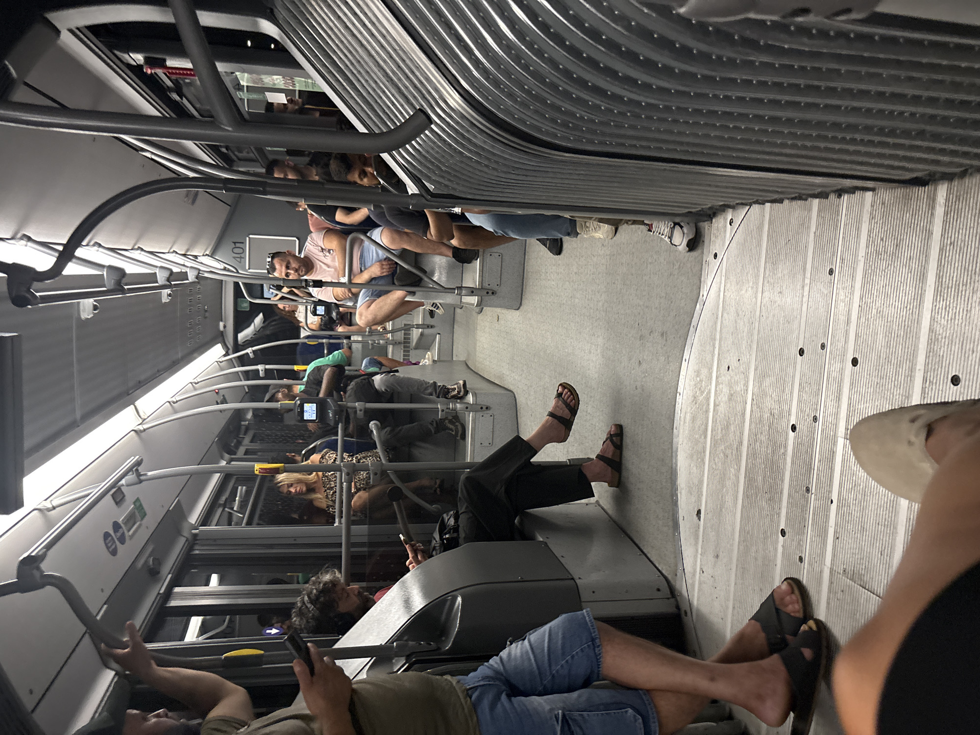 The image depicts the interior of a public bus in Venice, Italy, during what appears to be daytime. The bus is populated with several passengers, some seated and others standing. The lighting suggests it might be evening or a dimly lit interior, with artificial lights illuminating the space.
In the foreground, a person's legs are visible, wearing black shorts and black sandals with a brown strap. Their legs are crossed. To the left, another person, partially visible, is wearing denim shorts and dark sandals. This person is holding a smartphone in their left hand and appears to be standing.
Further inside the bus, a man with curly hair and headphones is seated, looking down at his phone. He is wearing dark pants and dark sandals. To his right, a woman with blonde hair, wearing a leopard print top, is seated. Adjacent to her, a man in a pink t-shirt and blue shorts is seated, looking forward.
The bus interior features rows of grey seats and metal poles for passengers to hold onto. The floor is made of a light-colored material with a ribbed texture, possibly wood or a composite. On the far right, a row of corrugated metal appears to be part of the bus's structure.
The overall scene conveys a sense of ordinary public transportation, with individuals engaged in their own activities. The presence of Venetian public transport suggests the location is within the city of Venice, Italy.