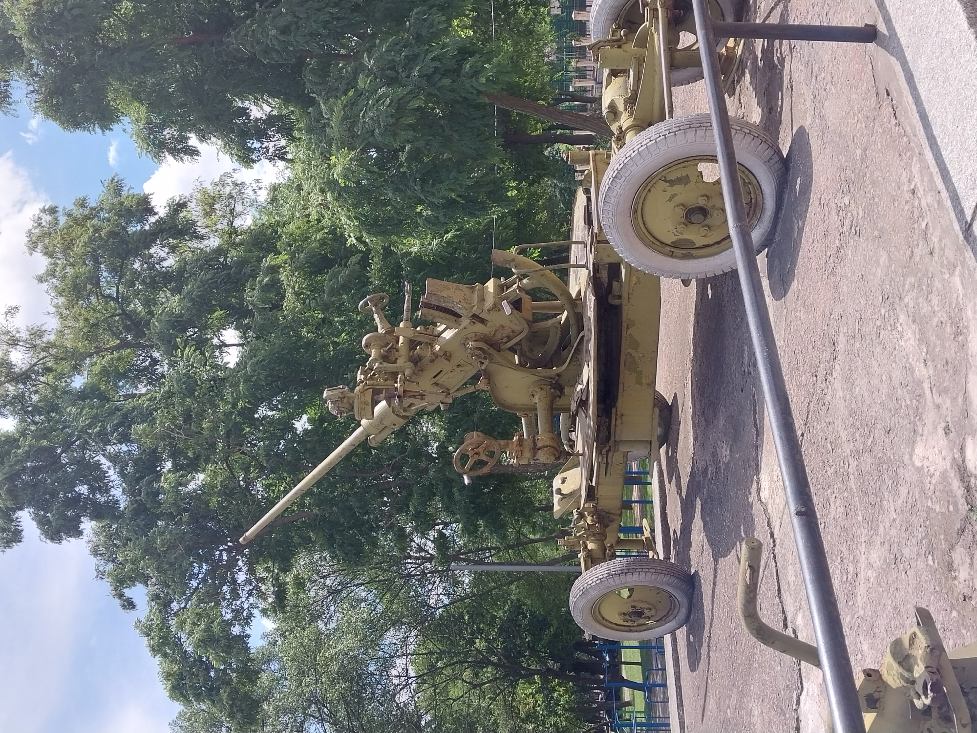 This is a full, slightly low-angle, eye-level shot of an anti-aircraft gun. The gun is painted olive drab and is mounted on a wheeled carriage. It is positioned on a concrete pad with cracked sections. The gun has a long barrel that angles upward and to the left. The carriage has two large, white-rimmed wheels visible on the left and right sides of the frame. The gun is a prominent object in the center and right of the image.
Behind the gun, a line of tall, green trees creates a dense backdrop with dappled sunlight filtering through. The trees are varied in height and species. Above the trees, a bright blue sky with scattered white clouds is visible.
In the foreground, a dark grey metal railing runs horizontally across the bottom of the image, obscuring the lower portion of the gun's carriage. Part of another metal pipe, also olive drab, is visible behind the railing on the left.
In the middle ground, beyond the trees, a blue metal fence is partially visible, suggesting a park or public area. Beyond the fence, a few dark shapes that might be parked vehicles are discernible.
The scene appears to be an outdoor exhibit or memorial, given the static display of the anti-aircraft gun and its placement in what seems to be a public space with trees and pathways. The overall impression is one of a sunny day in a green, urban environment. This artifact is located at the National Museum of the History of the Ukrainian Armed Forces in Dnipro, Ukraine, likely in the open-air exhibit area.