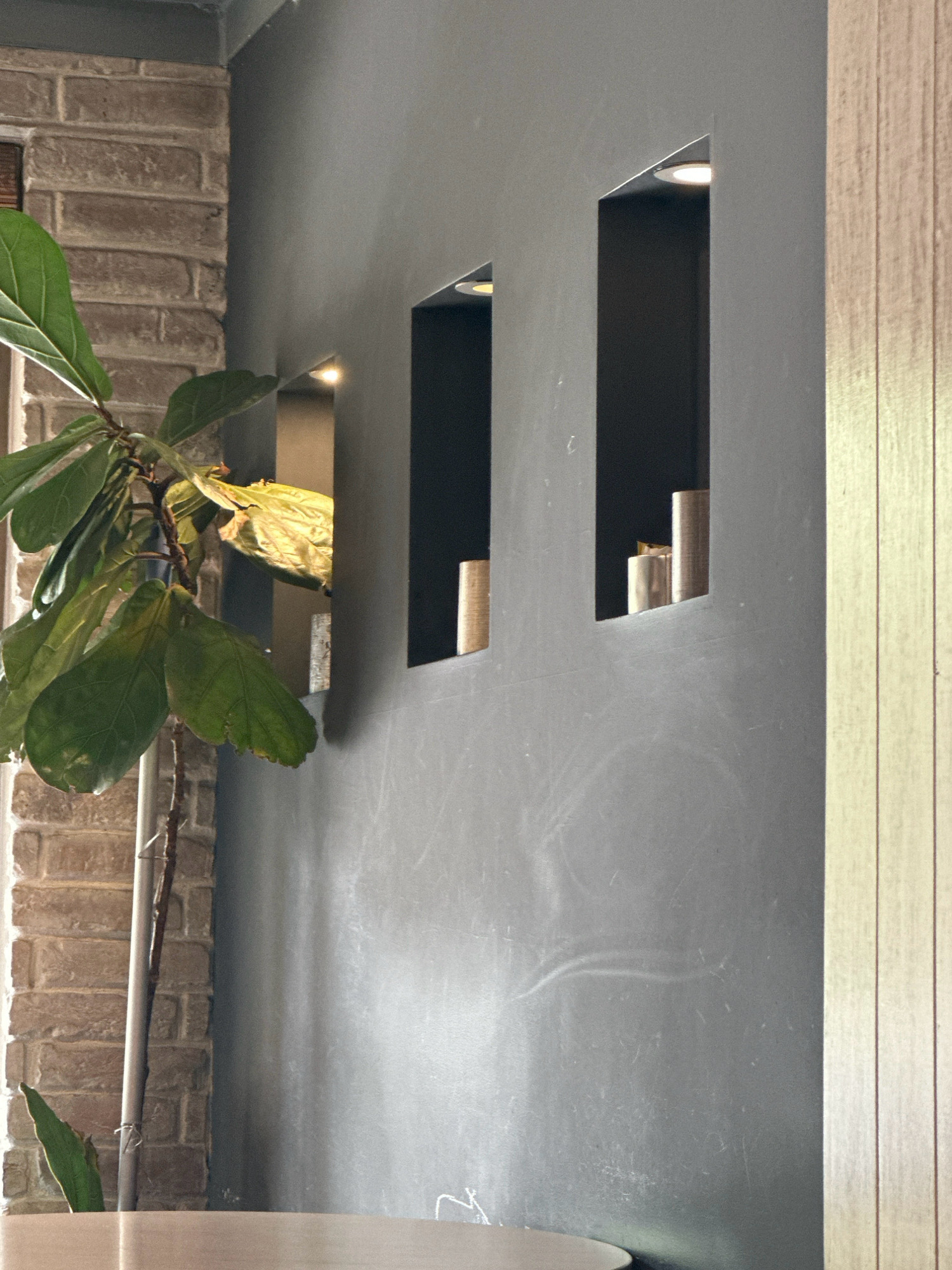 An indoor scene features a gray wall with two rectangular niches. Each niche contains cylindrical objects and is illuminated by a recessed light. To the left of the gray wall, a brick wall is visible, and a leafy plant with a thin stem grows in front of it. To the right of the gray wall, a light-colored wooden paneling extends vertically. The image does not contain any information to determine its location in Kathmandu Metropolitan City, Nepal.