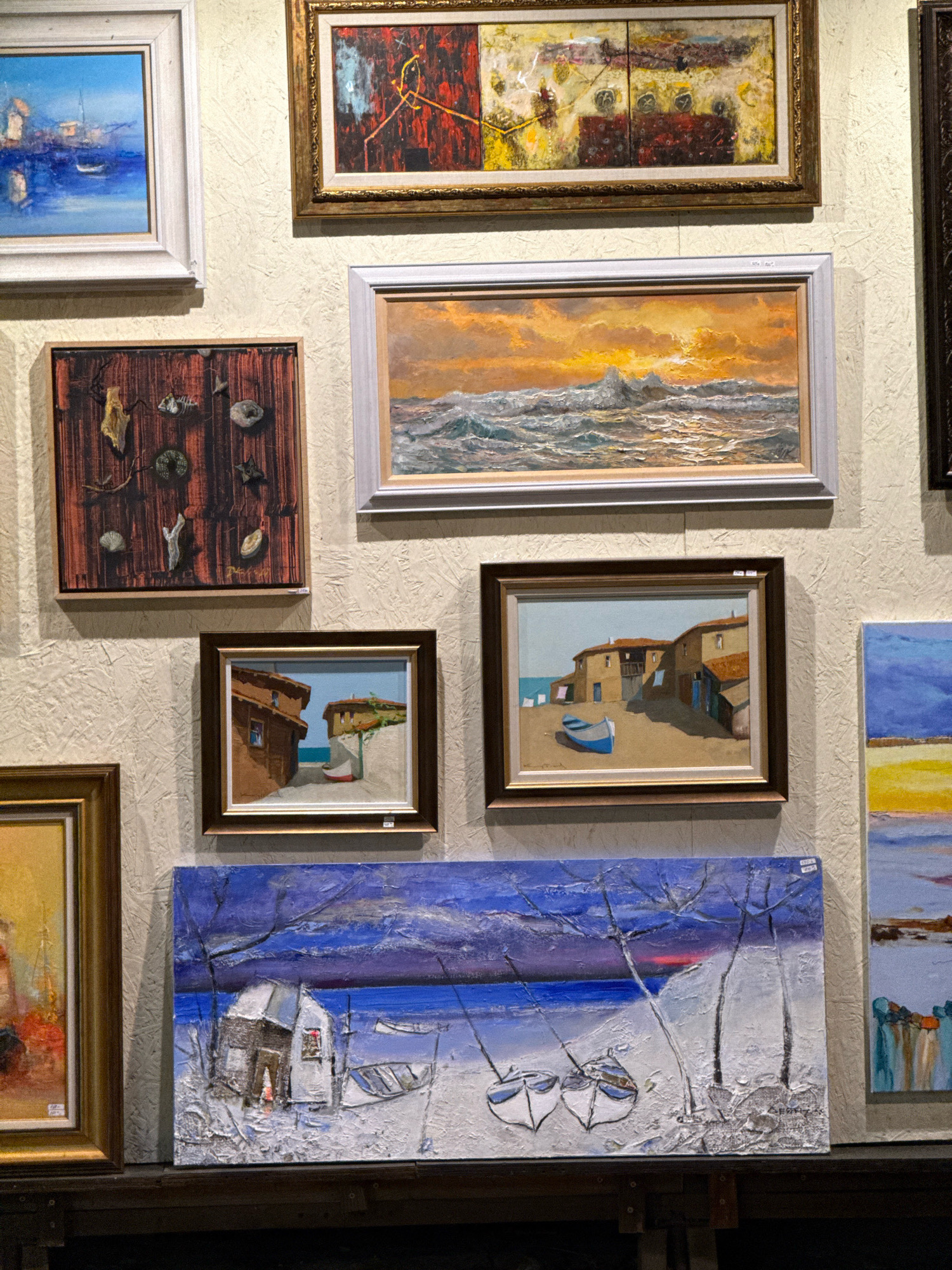 This is a photograph of several paintings displayed on a textured wall, likely in an art gallery or shop in Sozopol, Bulgaria. The paintings depict various scenes, primarily maritime or coastal.
Starting from the top left and moving clockwise:
* A framed painting of a bright blue sky over water, with a small white boat and distant buildings. The frame is light colored.
* To its right, a larger, horizontally oriented framed painting with a gold frame. This abstract work features bold strokes of red, black, yellow, and white, with some textured elements.
* Below the abstract painting and to the right of the first painting is another horizontally oriented framed painting. This artwork shows a dramatic sunset over a choppy sea, with a rocky island in the foreground. The frame is silver-toned.
* To the right of this, a portion of a vertically oriented painting is visible, featuring bands of color including blue, yellow, and brown, suggesting a landscape or seascape.
* Below the abstract painting and to the left of the sunset seascape is a framed painting of a Mediterranean village scene. Two brown buildings with red-tiled roofs and a small blue boat on a sandy shore are depicted. The frame is dark brown.
* Below the village scene and slightly to the left is a framed painting of a yellow and orange abstract or impressionistic landscape. The frame is gold.
* At the bottom, a large, horizontally oriented painting stretches across the frame. This textured artwork depicts a winter or snowy scene with a small, dilapidated building and two boats on a white, textured surface, possibly snow or ice. The background is a deep blue with hints of purple and red, suggesting a twilight sky. The frame is not clearly visible.
The wall itself has a rough, textured finish, appearing to be a light beige or off-white color. The lighting appears to be artificial, casting subtle shadows. The overall impression is a collection of art for sale or exhibition in a coastal town known for its artistic community and preserved old town architecture.