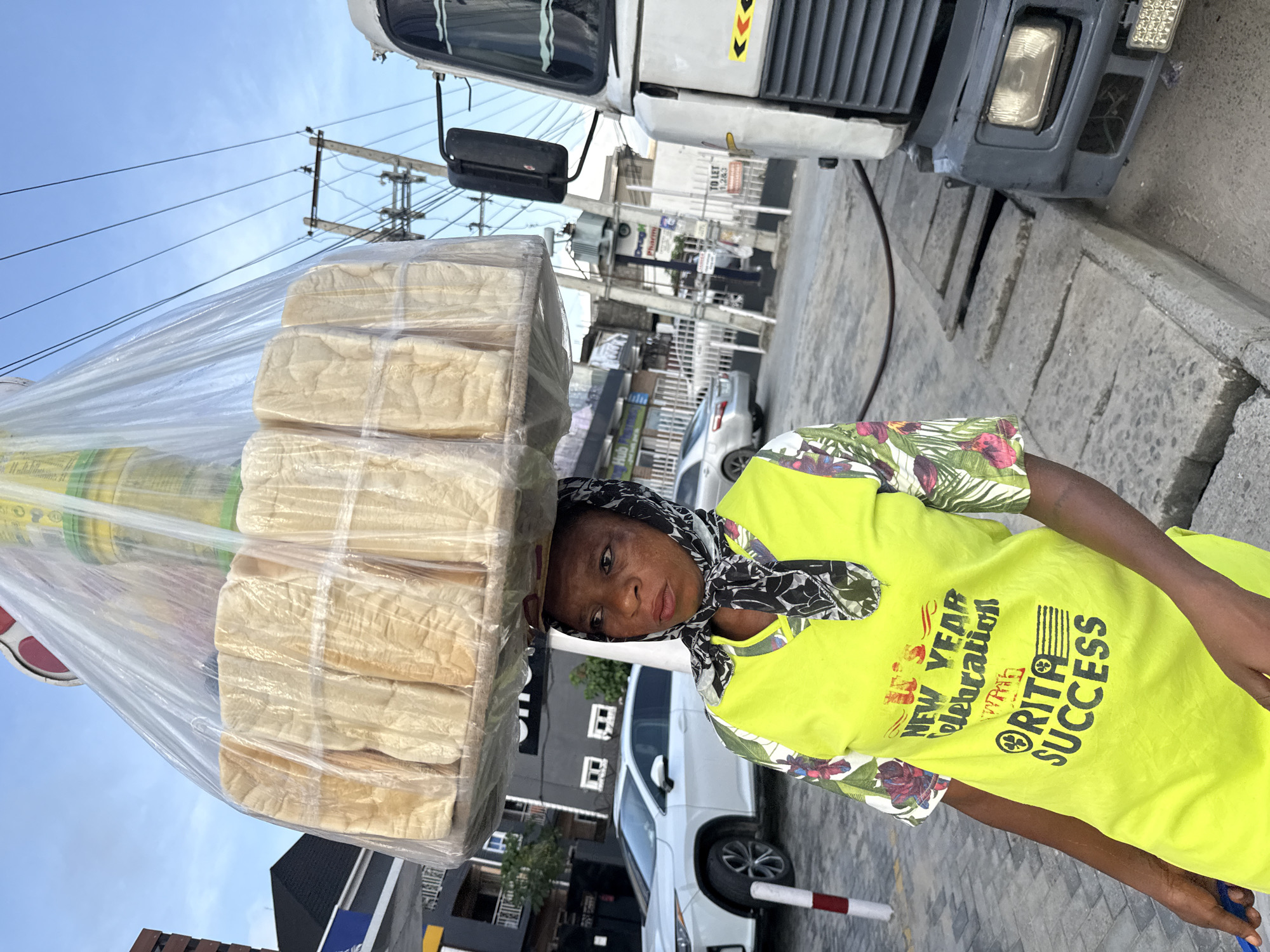 A medium, full-body outdoor shot captures a woman balancing a large stack of wrapped bread loaves on her head. She is positioned on what appears to be a city street in Ado, Nigeria.
She wears a bright yellow short-sleeved shirt with a floral pattern and a black-and-white patterned scarf tied around her head. Text and graphics are on her shirt: “NEW YEAR Celebration” and “RITA SUCCESS”. Her gaze is directed toward the camera.
The bread loaves are stacked high, wrapped in clear plastic, and rest directly on her head.
The environment includes urban elements like buildings, parked cars (a white Lexus and a gray car), street lighting, and power lines. A large white truck or van is parked nearby, partially visible in the upper portion of the frame. The pavement indicates that she is standing on a roadside or sidewalk.