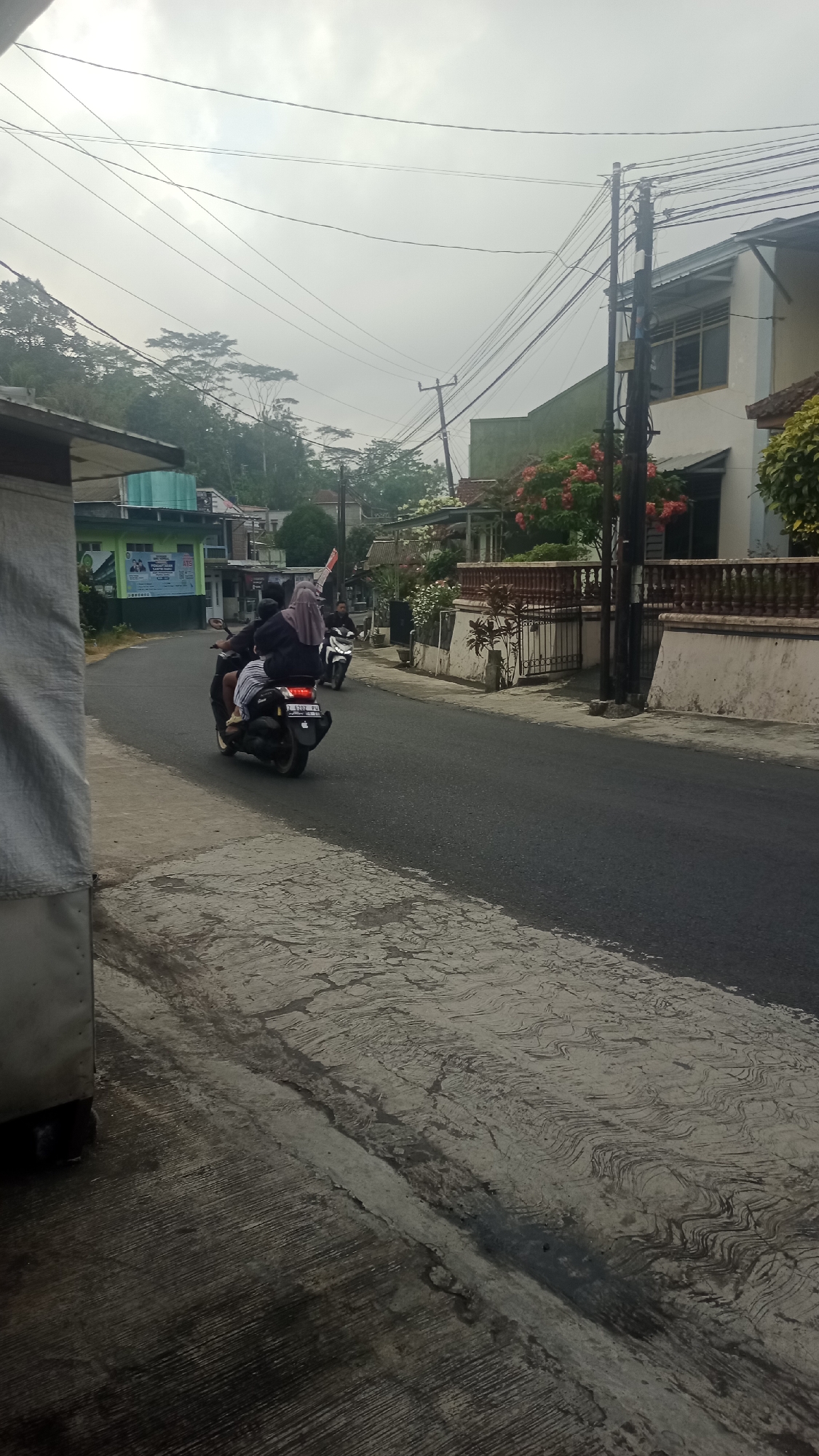 In the city of Karyamandala, Indonesia, two people are riding a motorcycle on a paved road. The person in the back is wearing a hijab and a striped skirt, and the person in front is wearing a black jacket. Another motorcycle with a single rider is visible further down the road. The road is bordered by a sidewalk with textured concrete. On the right side of the road, there is a building with a white and beige facade, a red-tiled roof, and a stone fence. Red and white flowers bloom in front of the building. On the left side of the road, there is a building with a green facade. Trees and utility poles with numerous wires are scattered throughout the scene. The sky is overcast and hazy.