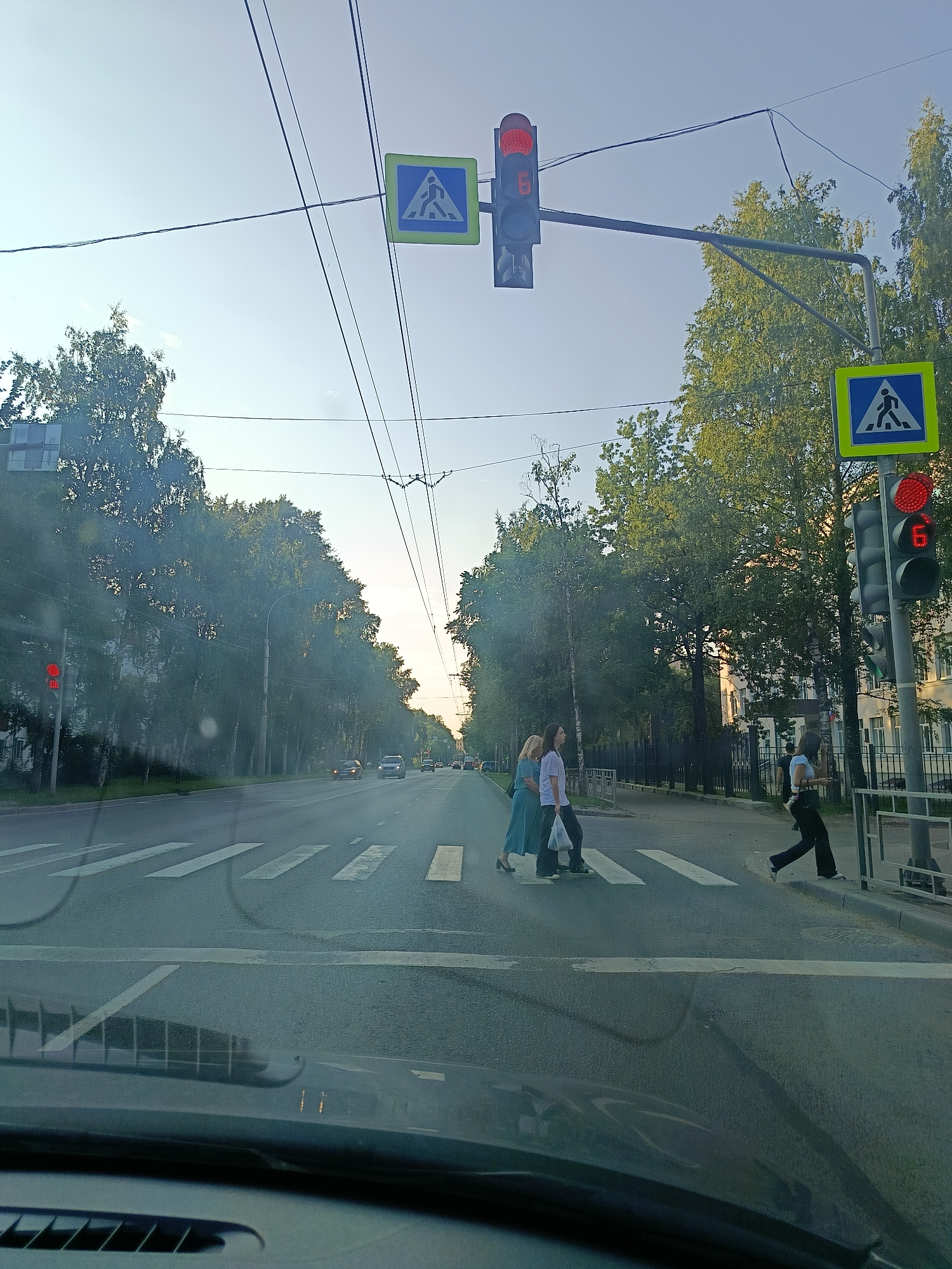 The image shows a view from a car of a street in Vologda, Russia. A pedestrian crossing is visible, with several people crossing the street. A traffic light is red, indicating that cars should stop. Trees line the street, and buildings are visible in the background. The sky is clear and blue, suggesting it is daytime. The image appears to have been taken in the late afternoon or early evening due to the angle of the sun and the long shadows.
