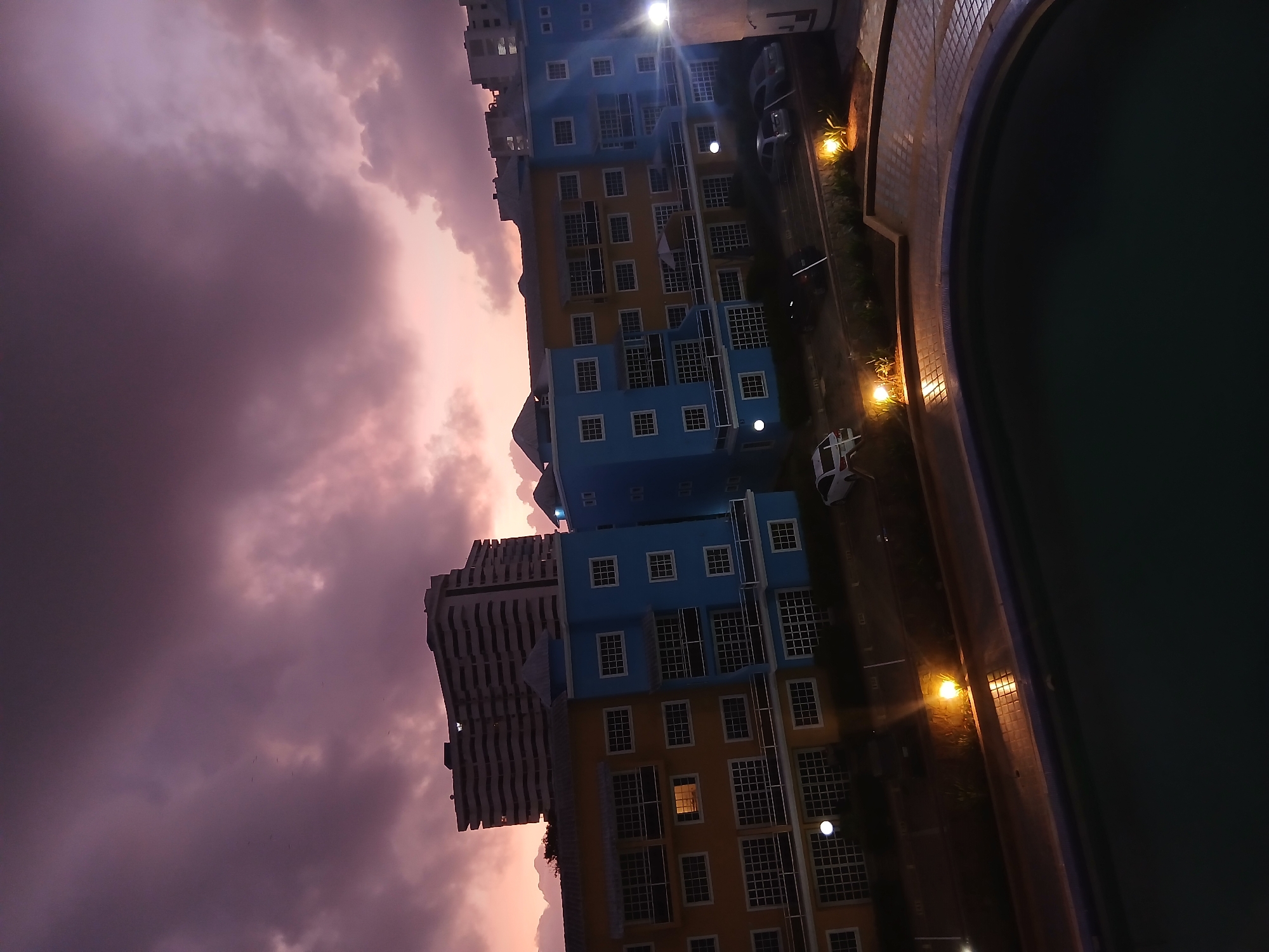 The image, oriented upright, depicts an urban or residential scene at twilight or early night, likely in Porlamar, Venezuela. The upper portion of the frame is dominated by a dramatic sky filled with large, dark clouds painted with shades of deep purple and soft pink, suggesting either the tail end of a vibrant sunset or an impending weather event.
In the mid-ground, a distinctive multi-story building features a facade composed of alternating sections of bright blue and yellow, punctuated by numerous grid-patterned windows. Many of these windows glow with internal lights. To its left, a taller, grey building with a textured, ribbed architectural design is partially visible, silhouetted against the sky.
The foreground on the right side features a dark, still swimming pool, its calm surface reflecting the surrounding light. A paved area or walkway separates the pool from the main building. This path is illuminated by several warm, yellow artificial lights positioned at ground level, which highlight small landscaped sections with foliage. A very bright, overhead streetlight emits an intense glow from above the blue and yellow building, creating a prominent starburst. A few indistinct vehicles are visible on the paved surface, with part of a white car on the far right edge.
The scene is devoid of people, presenting a quiet moment. The contrasting elements of the atmospheric, colorful sky, the brightly lit building, and the calm, dark pool contribute to a mood that feels both serene and dramatically lit by the interplay of natural dusk light and artificial illumination. No discernible text is visible.