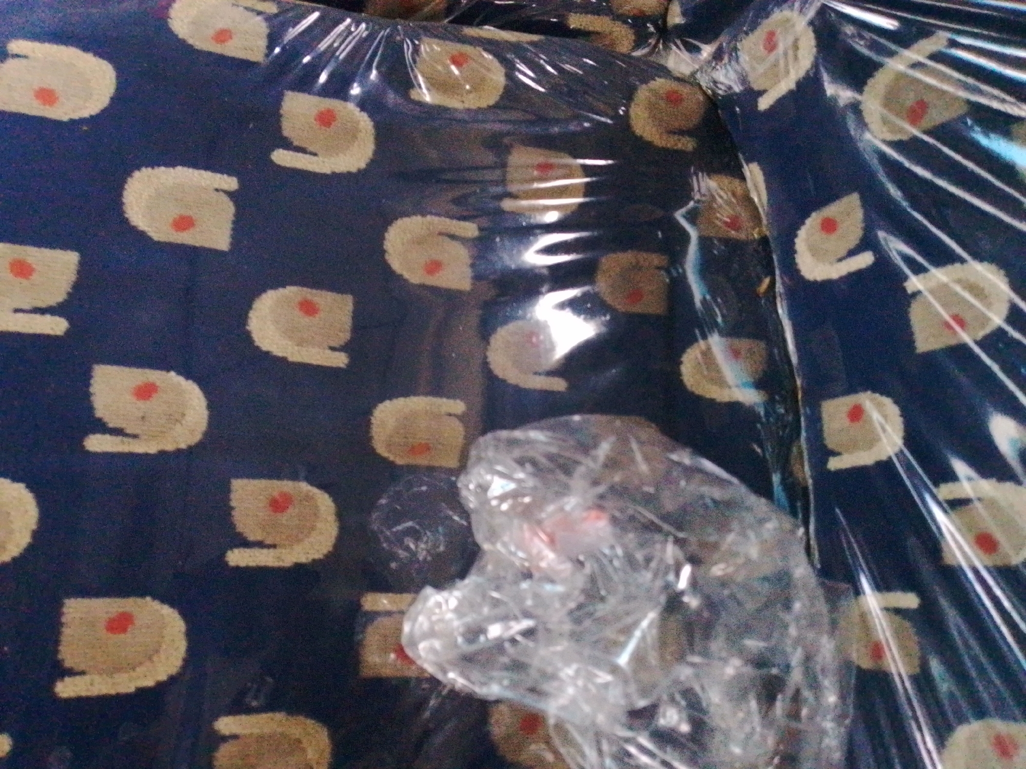 The image shows a close-up of a dark blue fabric with a repeating pattern of light beige shapes, each containing a small red circle in the center. The fabric is covered in clear plastic sheeting, which reflects light and distorts the view. In the foreground, a crumpled piece of clear plastic sheeting obscures a portion of the fabric. The scene appears to be indoors, possibly in a storage area or during transit, as suggested by the plastic wrapping. The specific location of Barikin Jirga Summa, Nigeria, cannot be determined from the visual information provided in the image. There are no people or specific actions depicted.