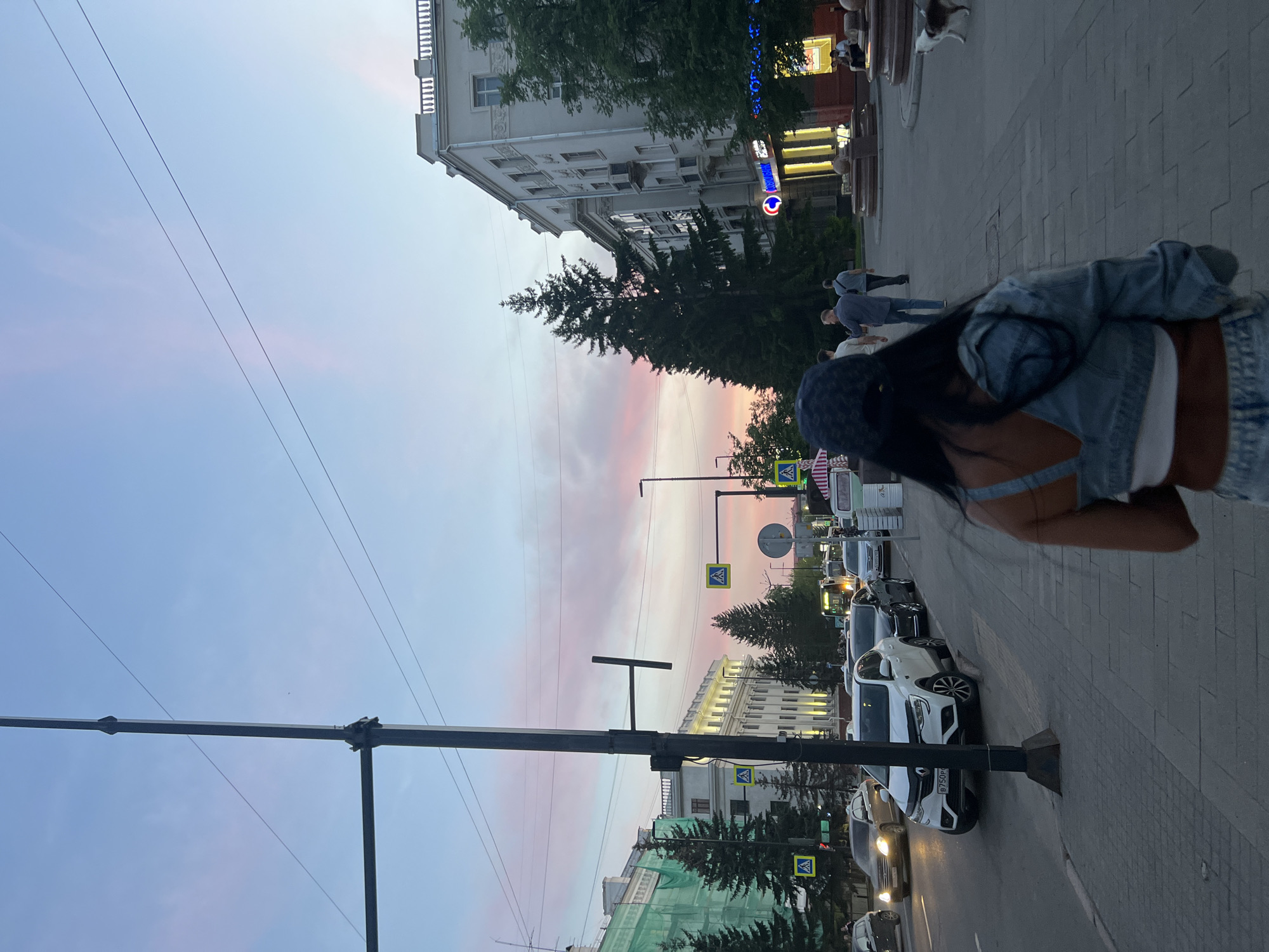 The image depicts an evening scene on a city street in Krasnoyarsk, Russia, captured during twilight. The sky is a prominent feature, displaying a beautiful gradient of soft pink near the horizon transitioning to a lighter blue overhead, traversed by multiple power lines.
In the foreground, a person wearing a denim jacket and a dark cap is seen from behind, walking along a wide, paved sidewalk. To their left, the street is bustling with activity. Parked cars, including a white Renault displaying the Russian license plate "B750PT," line the roadside, while other vehicles are visible in motion.
The street is flanked by multi-story buildings of varying architectural styles, some featuring ornate facades. A large blue neon sign with the "ВТБ" (VTB) bank logo is clearly visible on one of the buildings, adding to the urban ambiance. Clusters of trees, primarily conifers, are interspersed between the buildings and along the street, providing greenery. Several yellow pedestrian crossing signs are visible. Further along the sidewalk, other pedestrians can be seen, with one person walking a small dog near what appears to be a stone monument or fountain. The artificial lights from buildings and vehicles are beginning to glow, signaling the approach of night in this active urban setting.