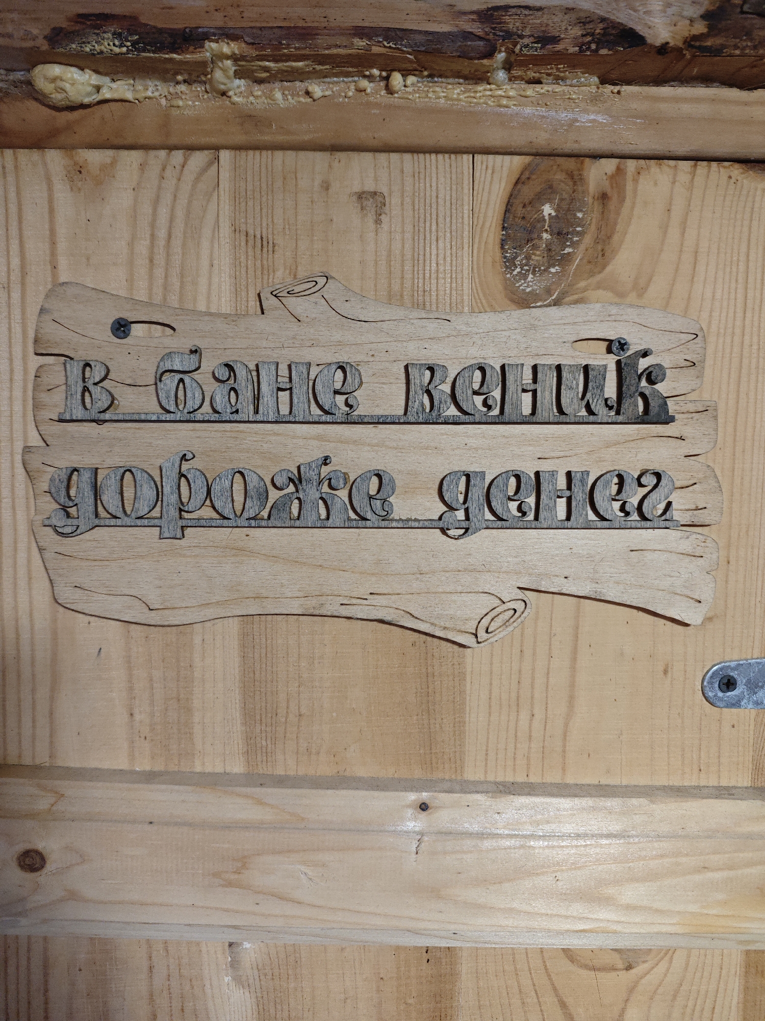 The image shows a wooden sign hanging on a wooden wall. The sign is shaped irregularly, like a piece of wood with stylized knots. It is inscribed in Cyrillic text in a decorative font. The first line reads "В бане веник," and the second line reads "дороже денег." The text is made of a contrasting material, possibly wood or metal, attached to the base of the sign. Two screws are visible, securing the sign to the wall. The wooden wall consists of planks with visible wood grain and knots. There's some sealant or insulation material at the top edge where the wall meets the ceiling. A metal hasp or latch is visible to the right of the sign. There is no visible information to confirm the location within Фоки, Russia.