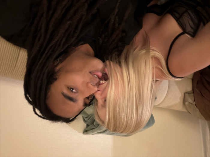 A man with long dreadlocks and a woman with blonde hair lie on a sofa together, their faces close and touching. The woman wears a black lingerie top. The man has a black zip-up shirt or jacket. They appear to be intimate. The scene is casually lit.