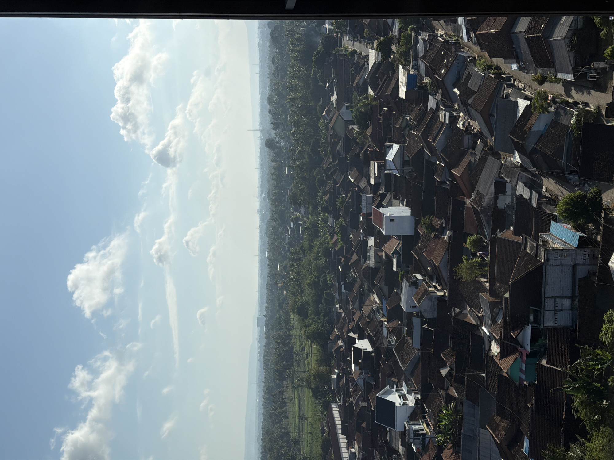 This is a high-angle aerial photograph of a dense residential area in Dadapan, Indonesia, viewed from a building. The scene is dominated by a vast expanse of rooftops, predominantly in shades of terracotta and grey, indicating a densely populated urban or semi-urban environment. The houses are closely packed, with narrow streets winding through the settlement. Greenery, consisting of trees and what appear to be agricultural fields, is visible in the background, suggesting the area is surrounded by or interspersed with natural landscapes. The sky above is a clear blue, with scattered white cumulus clouds, indicating fair weather. The overall impression is that of a typical Southeast Asian town or city, characterized by its extensive roof coverage and the integration of residential areas with surrounding vegetation.