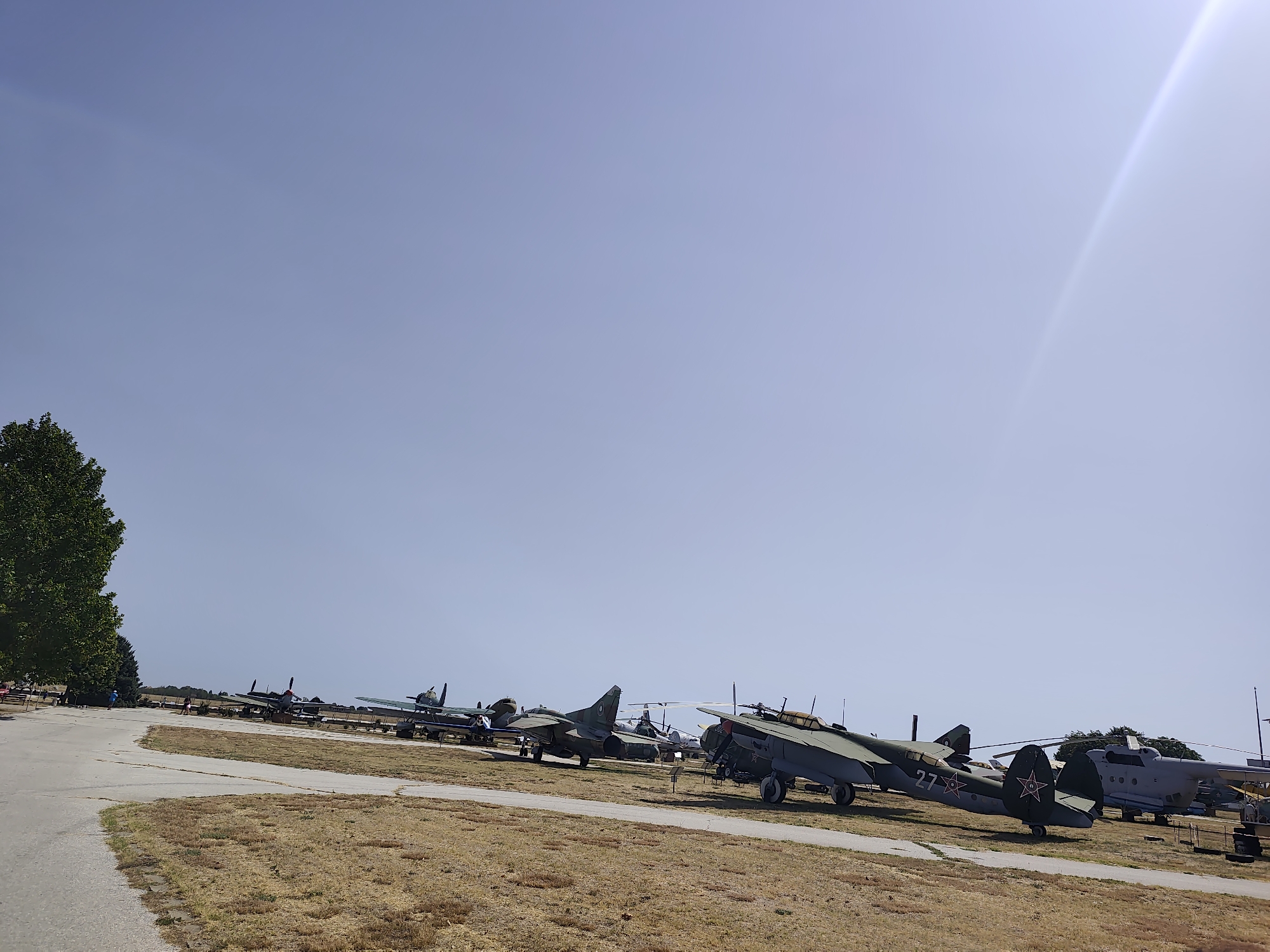 The image shows a clear sky with a bright sun glare on the right side. In the foreground, a paved walkway leads to a dry, grassy field. Several vintage aircraft are parked on the field in a line, appearing to be part of an aviation museum or exhibition. The aircraft are a mix of propeller-driven planes and at least one helicopter. Some of the planes are painted in military camouflage. In the background, a line of trees can be seen, and further back, there are some buildings or structures. A few people are visible walking on the walkway and near the aircraft. The location is identified as Krumovo, Bulgaria.
