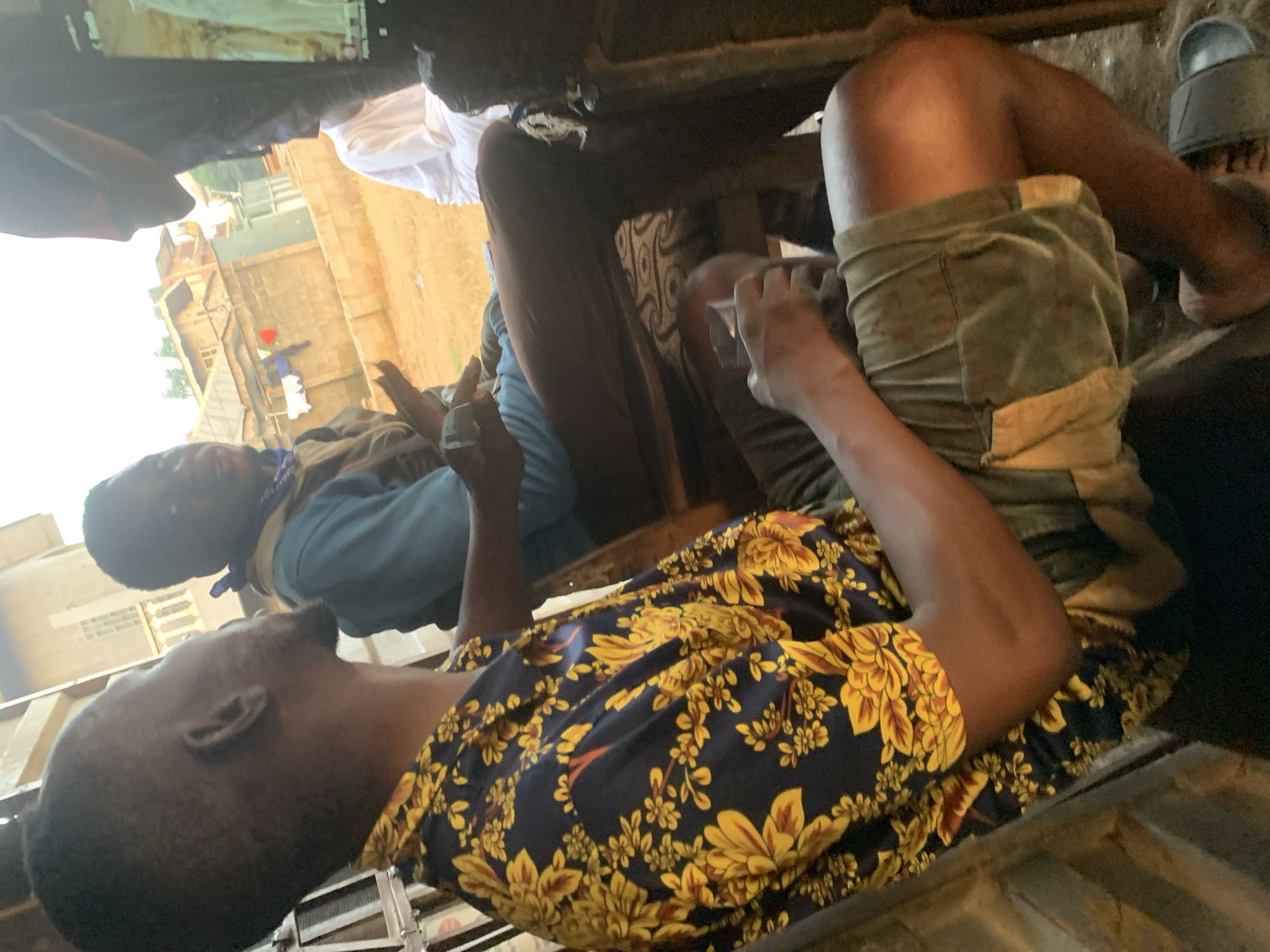 The image depicts a group of individuals gathered outdoors in what appears to be a residential or commercial area in Shagamu, Nigeria. In the foreground, a man wearing a dark blue shirt with a vibrant yellow floral pattern is seated, facing away from the camera. He is wearing olive-green shorts. Beside him, another man in a blue t-shirt and a brown vest is also seated. A third individual, dressed in a white garment, is partially visible to the right. The background shows buildings with corrugated metal roofs and concrete walls, suggesting an urban or semi-urban setting. Clothes are hung out to dry on a line in the middle ground. The ground is covered in dirt and debris. One of the men is holding a clear glass. The lighting suggests it is daytime, possibly late afternoon, with warm tones casting shadows. The overall scene conveys a casual gathering in a community setting.